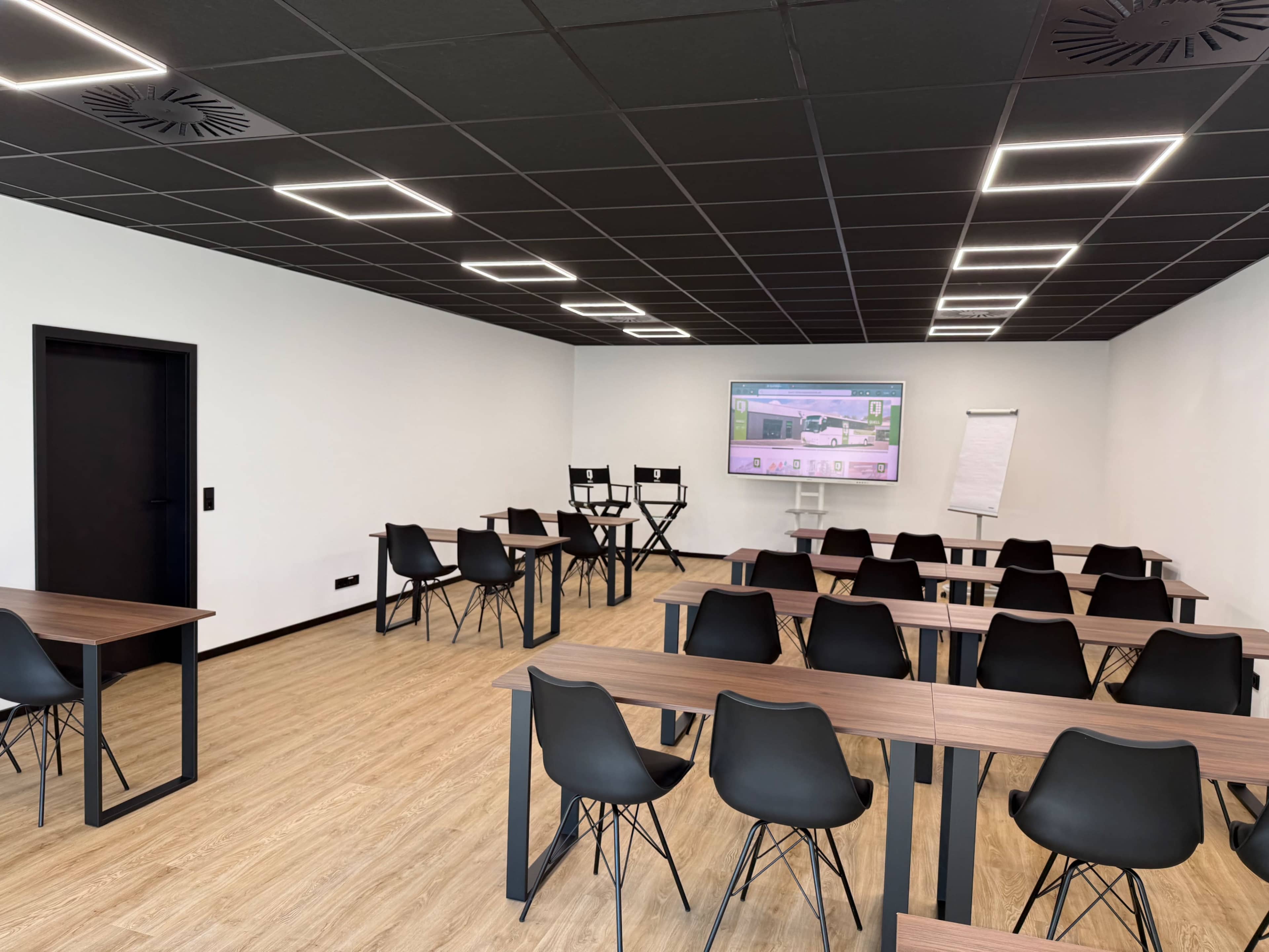 The image shows a modern classroom with rows of desks and chairs, a large screen at one end, and a flip chart against the wall.