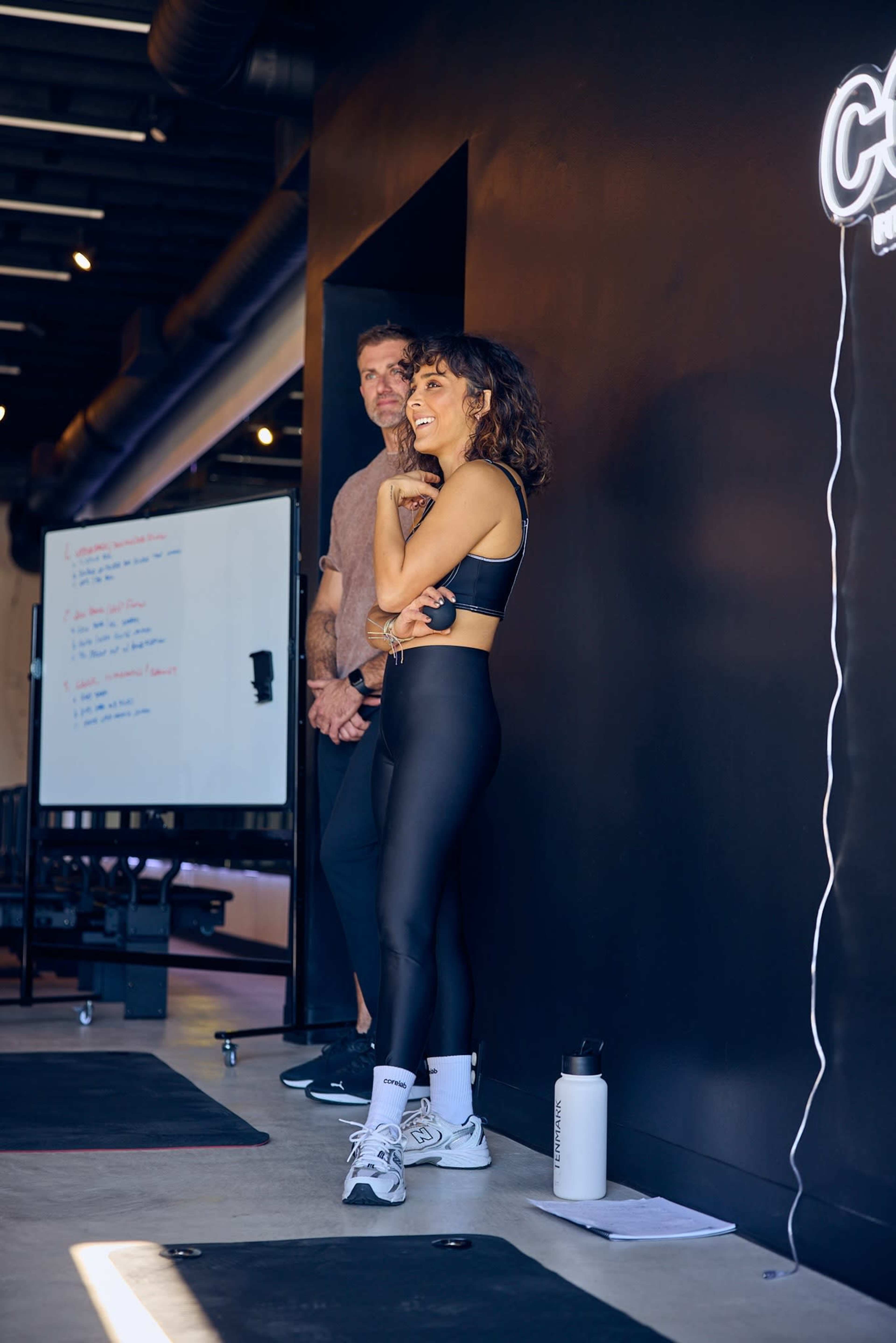 A woman in athletic wear stands smiling while a man in a gray shirt watches in a gym setting with a whiteboard in the background.