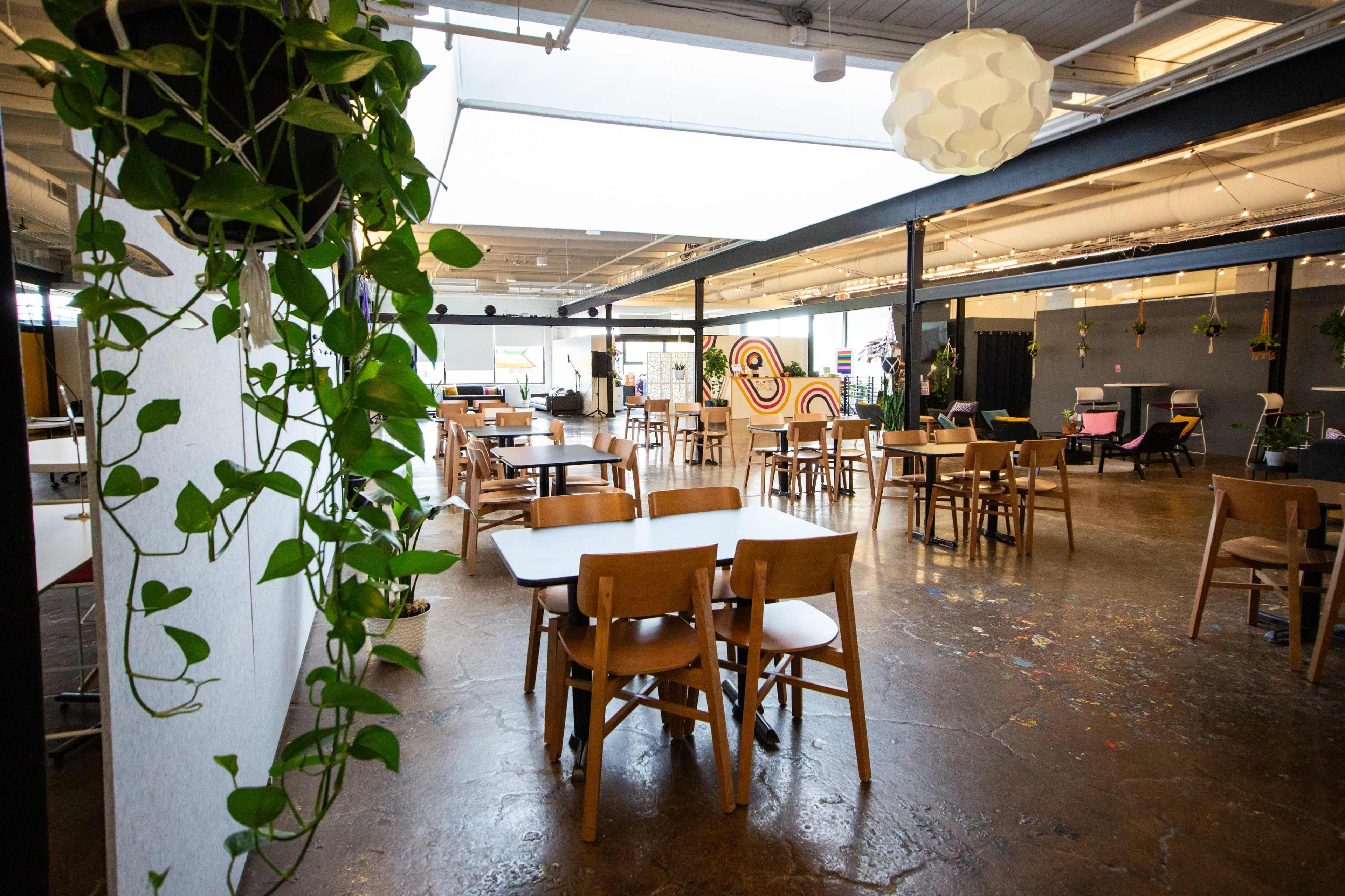 The image shows a spacious indoor area with wooden tables and chairs, hanging plants, and modern decor elements.