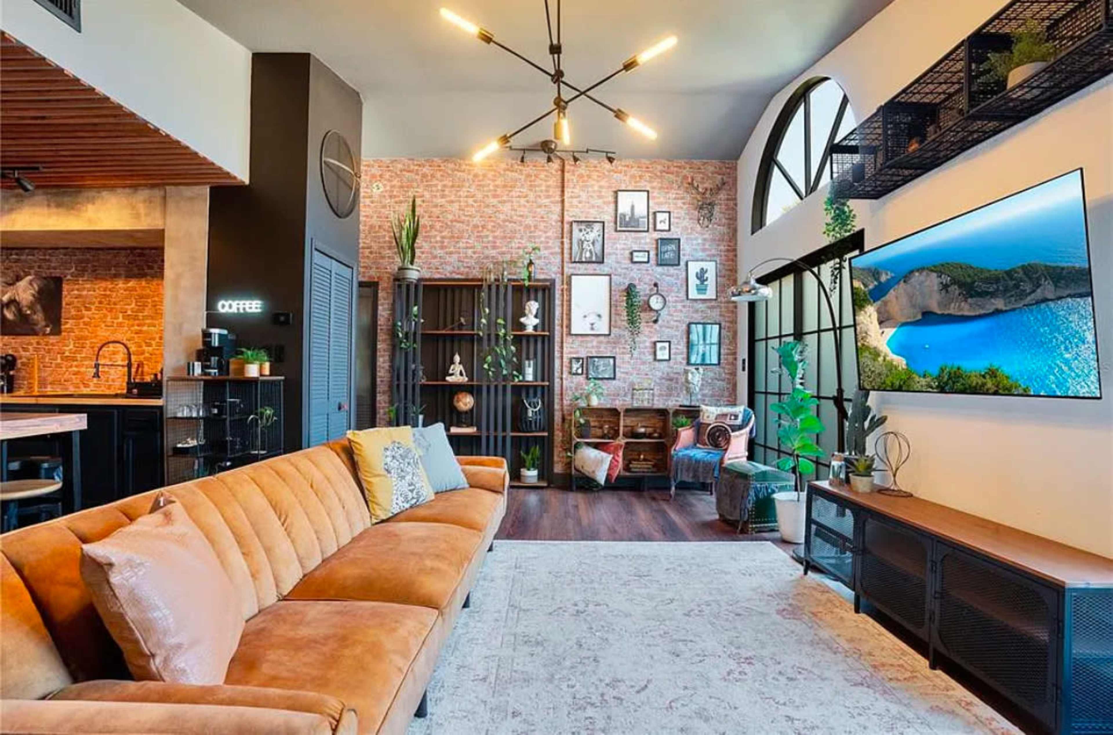 A modern living room features a tan sofa, a large TV mounted on a brick wall, and various decorative elements on shelves.