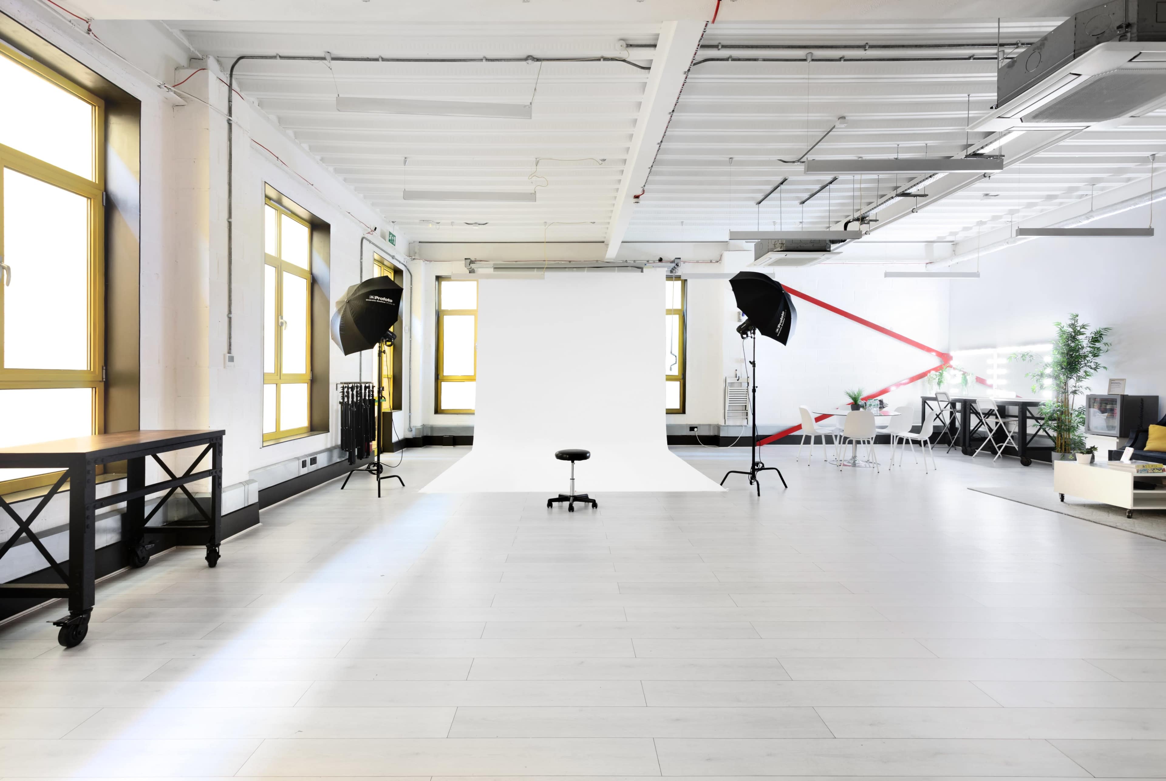 The image shows a spacious photography studio with a white backdrop, two light umbrellas, and modern furniture arranged in the background.