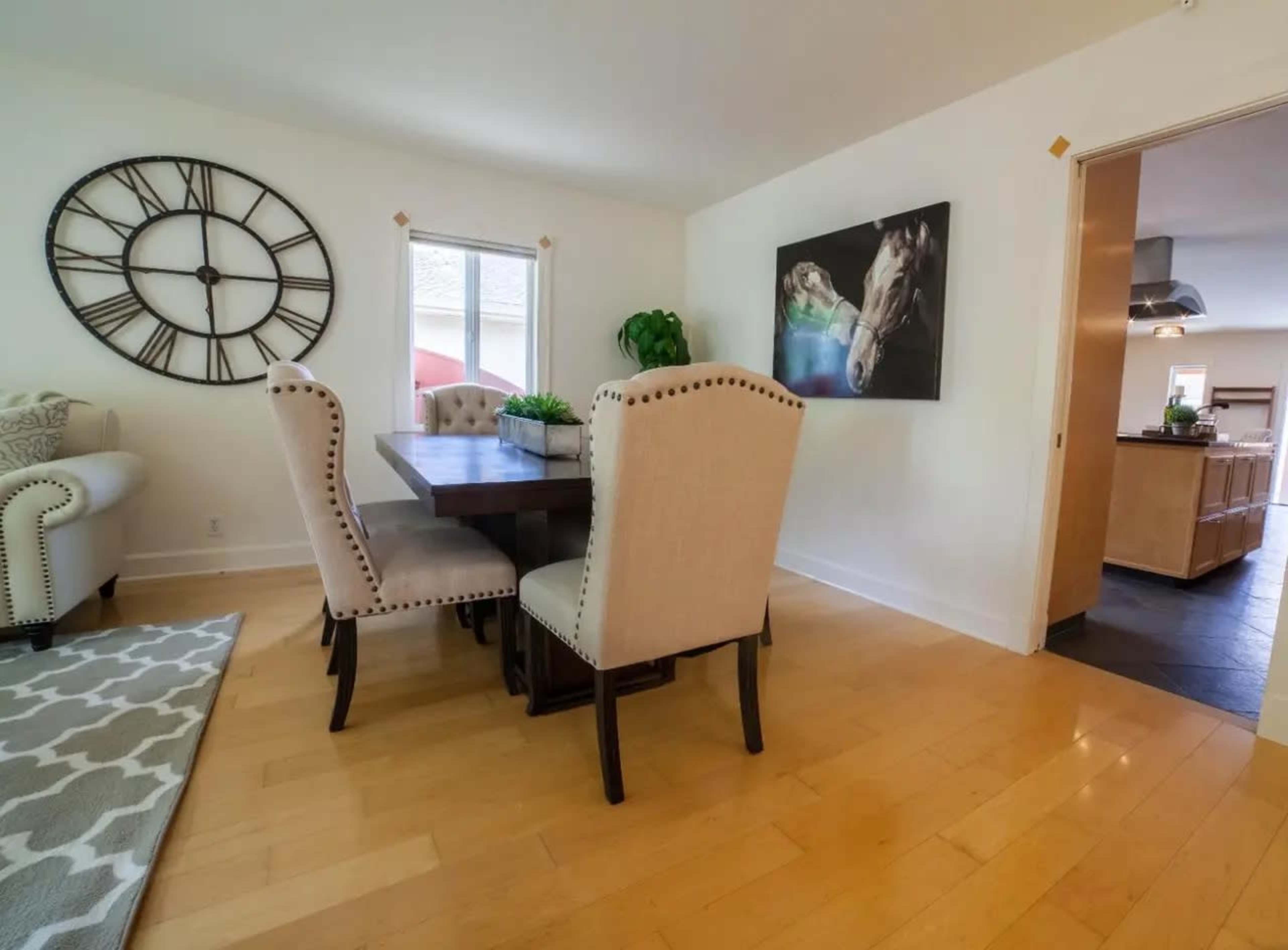 A dining area features a wooden table surrounded by four upholstered chairs, with a large wall clock and a painting of a horse on the adjacent wall.