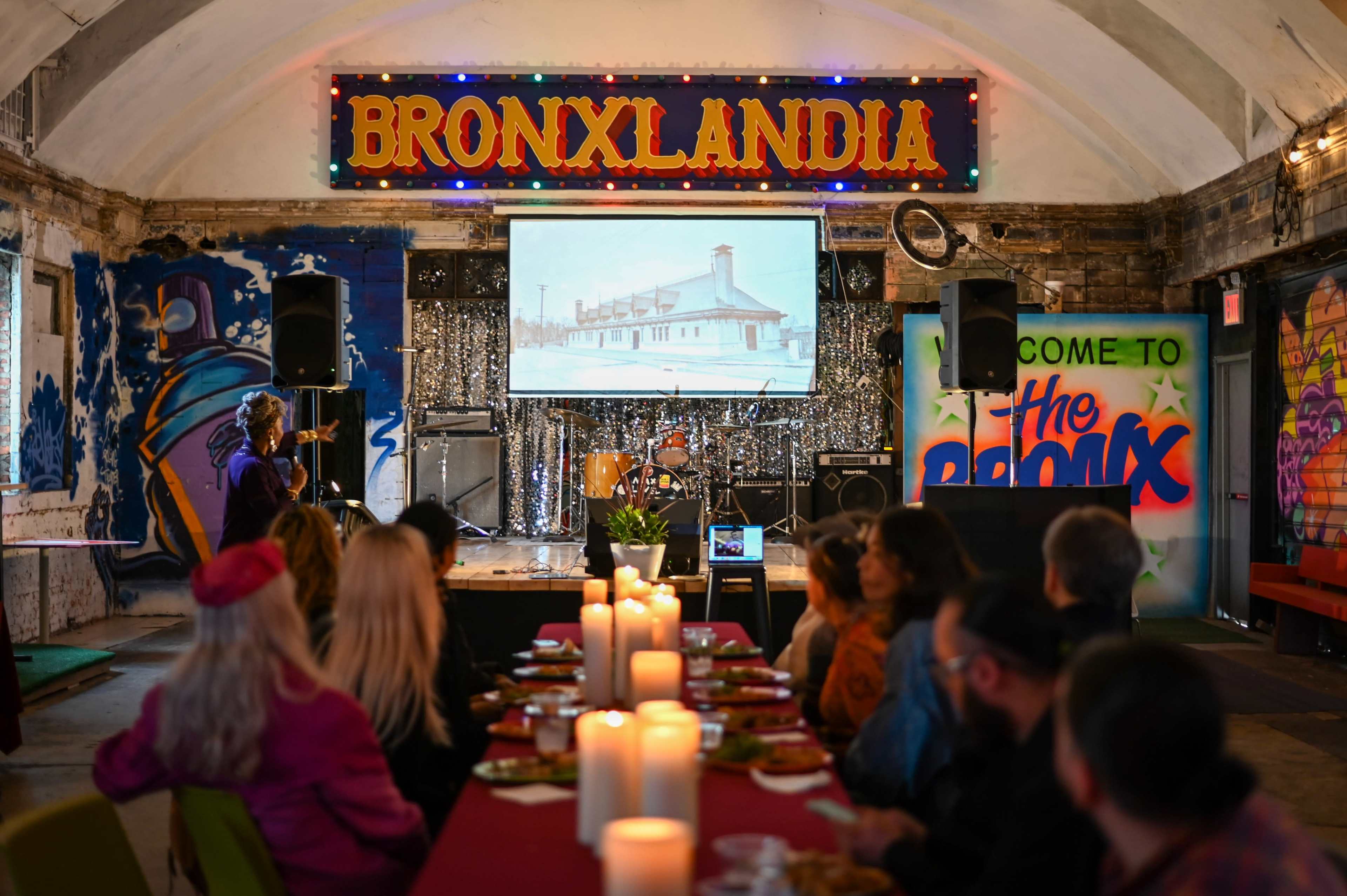 A group of people sits around a long table decorated with candles and food, while a presentation is projected on a screen in a colorful venue titled "BronXlandia."