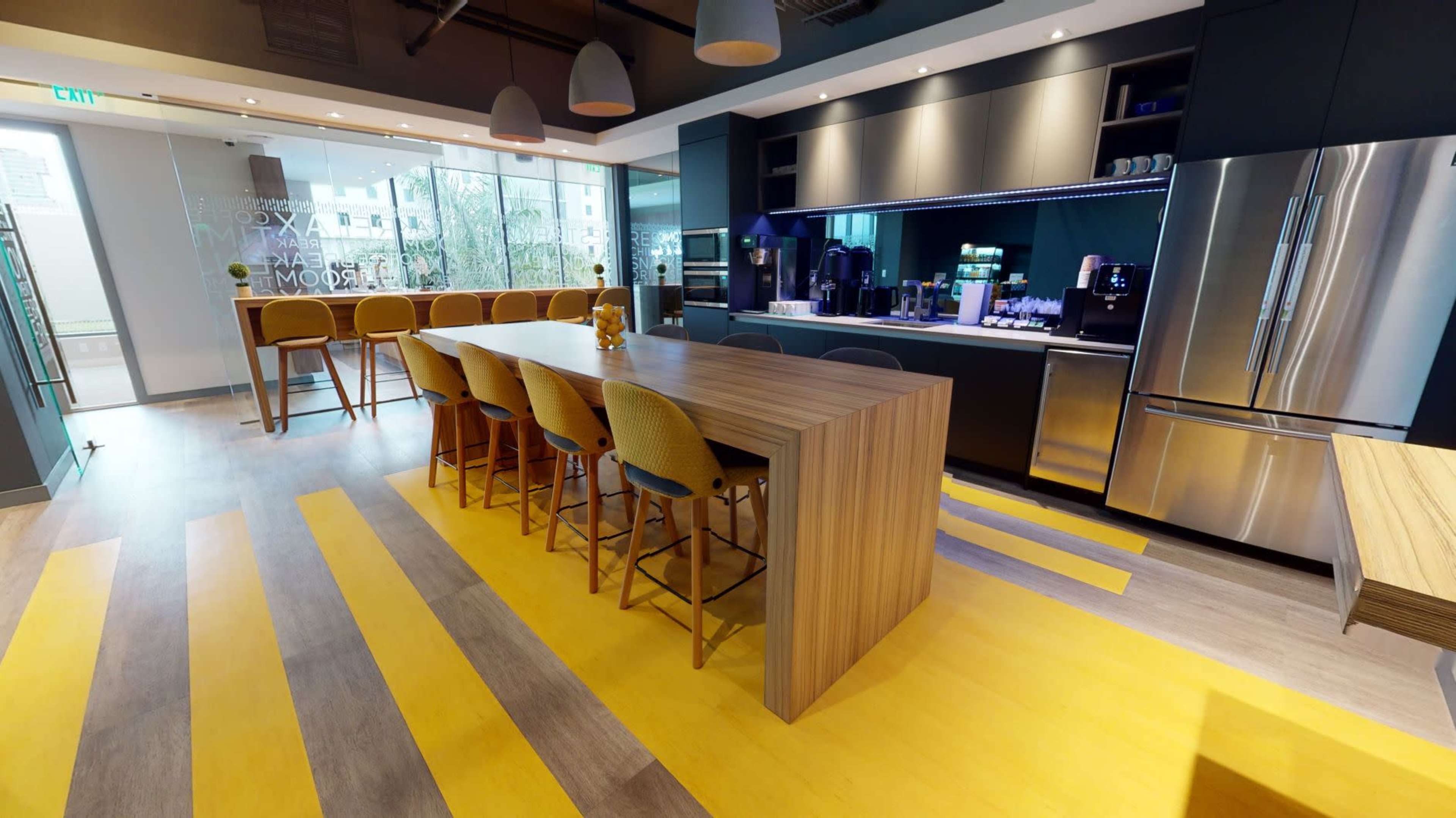 A modern kitchen area features a long wooden island with bar stools, stainless steel appliances, and large windows allowing natural light.