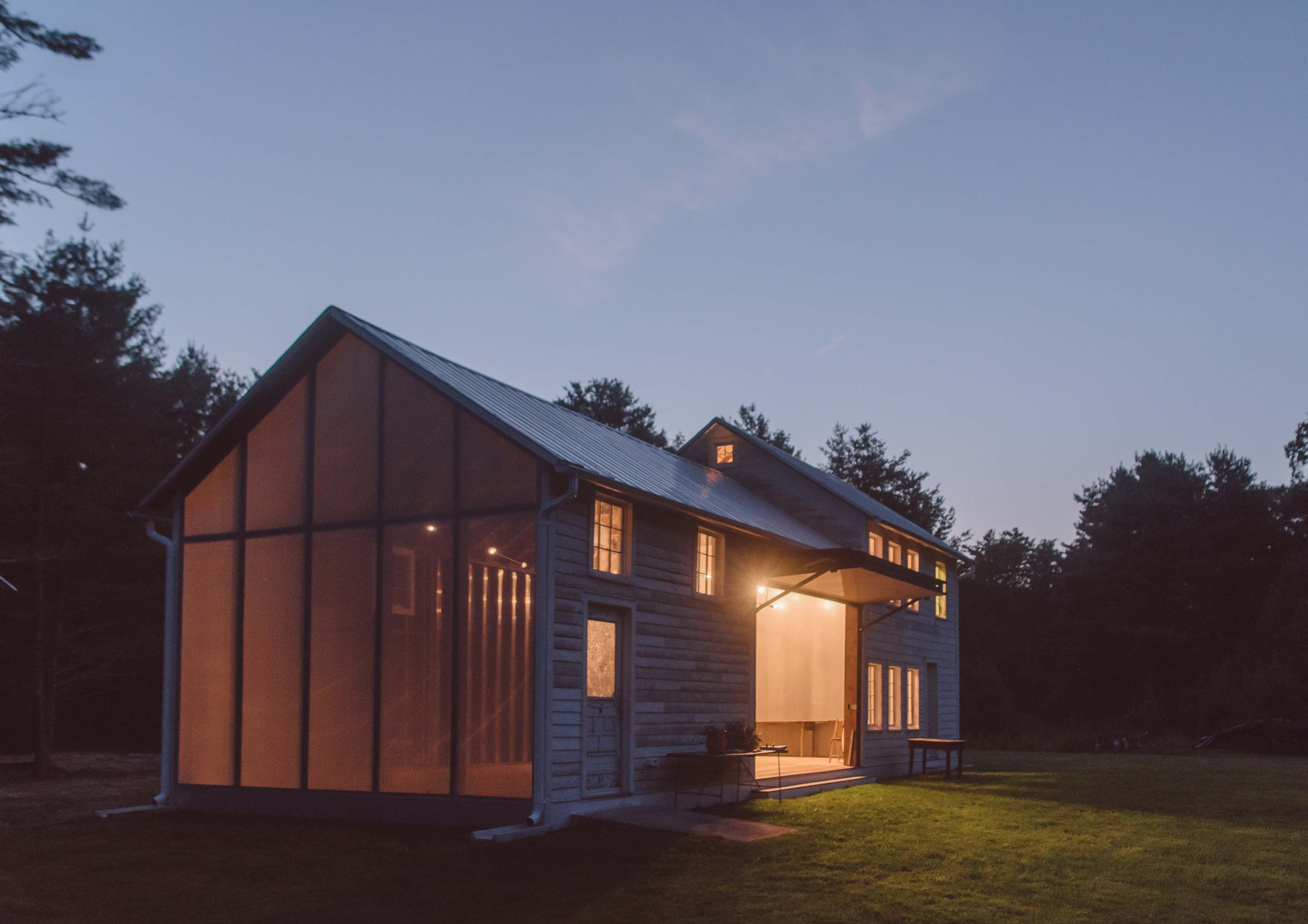 A modern, two-story house with large windows and a screened porch is illuminated at dusk, surrounded by trees.