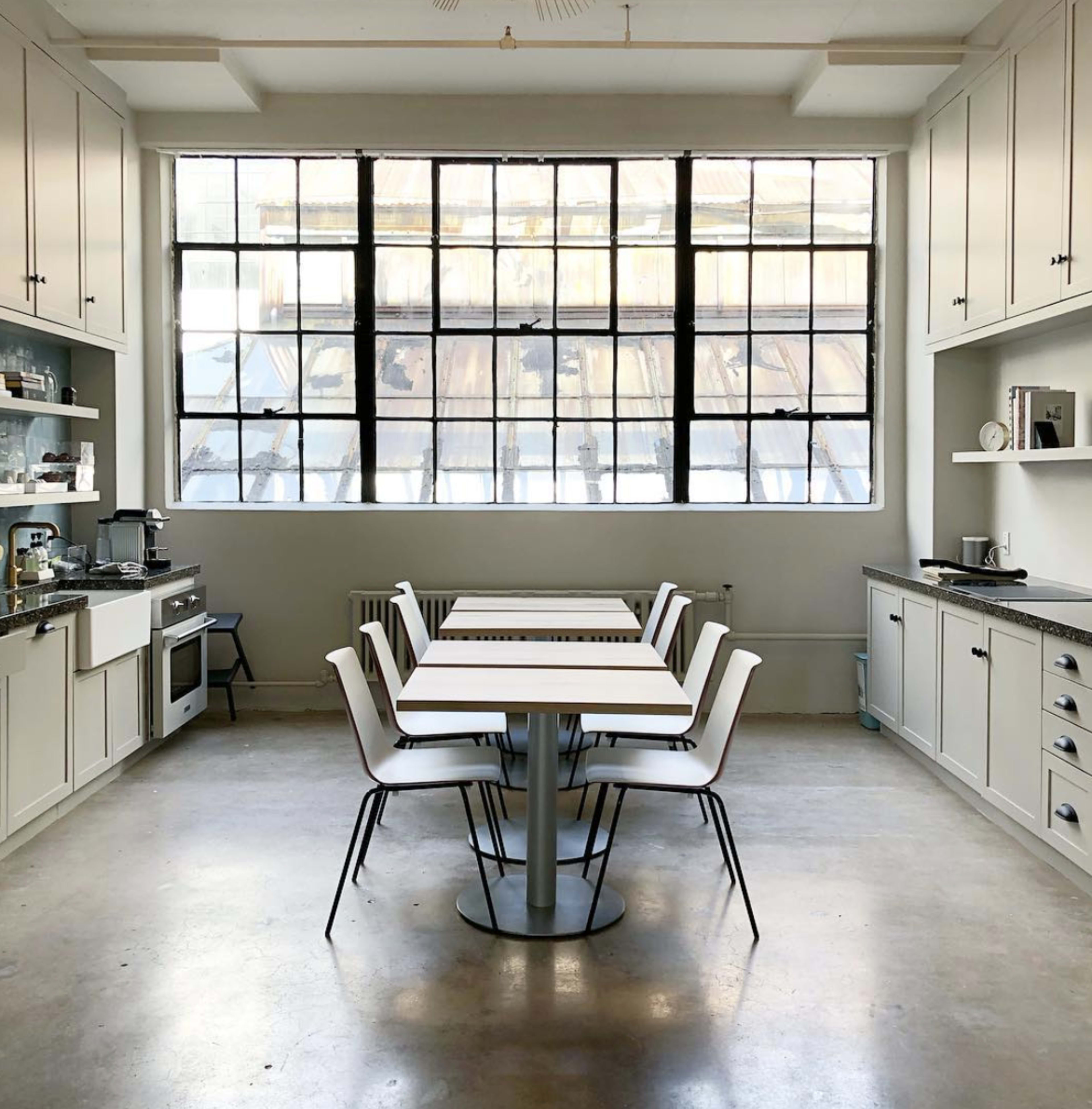 A modern kitchen features a central dining table surrounded by chairs, with large windows allowing natural light to fill the space.