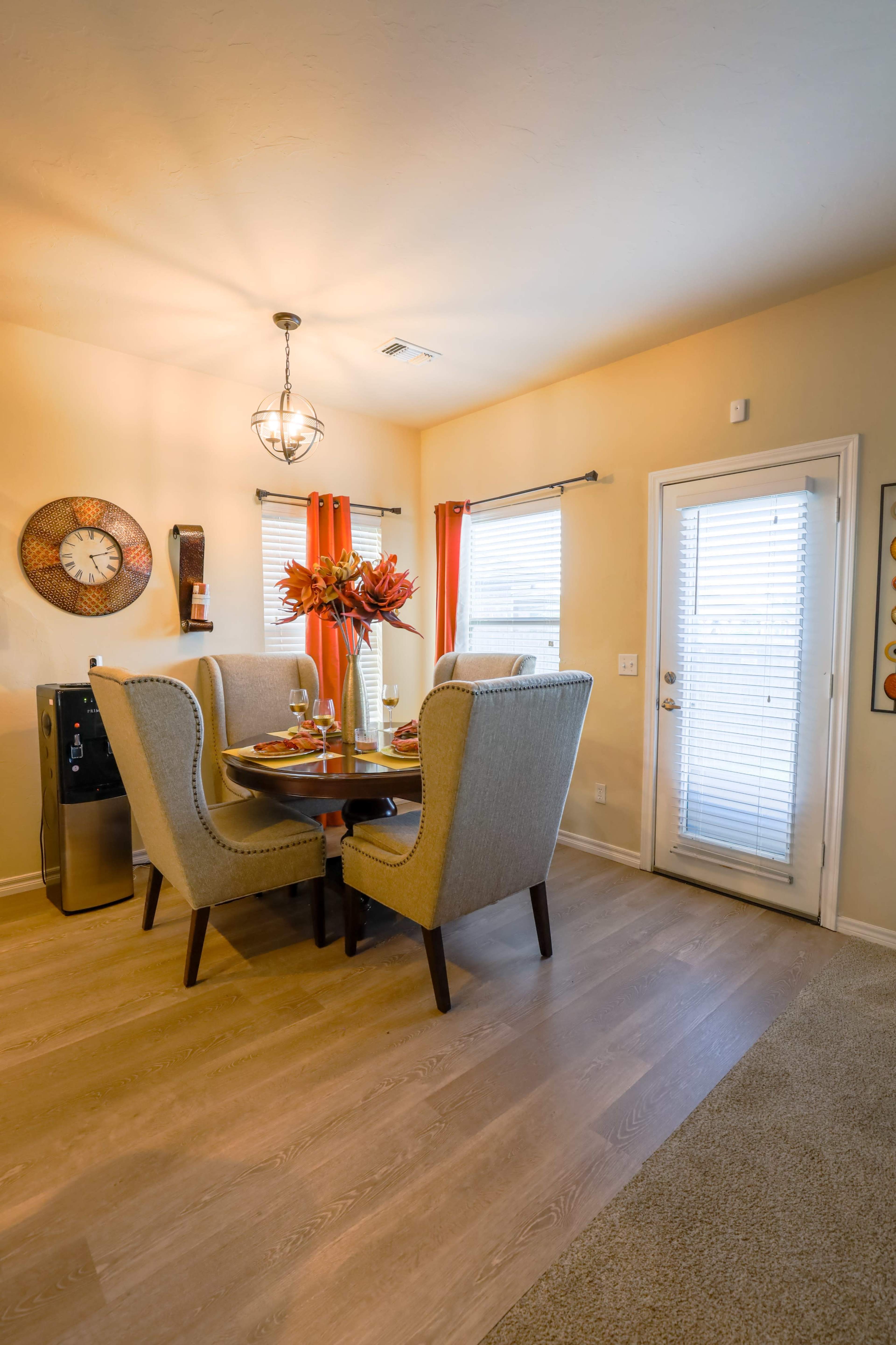 A dining area features a round table surrounded by four upholstered chairs, with window treatments that include orange curtains and a door leading outside.