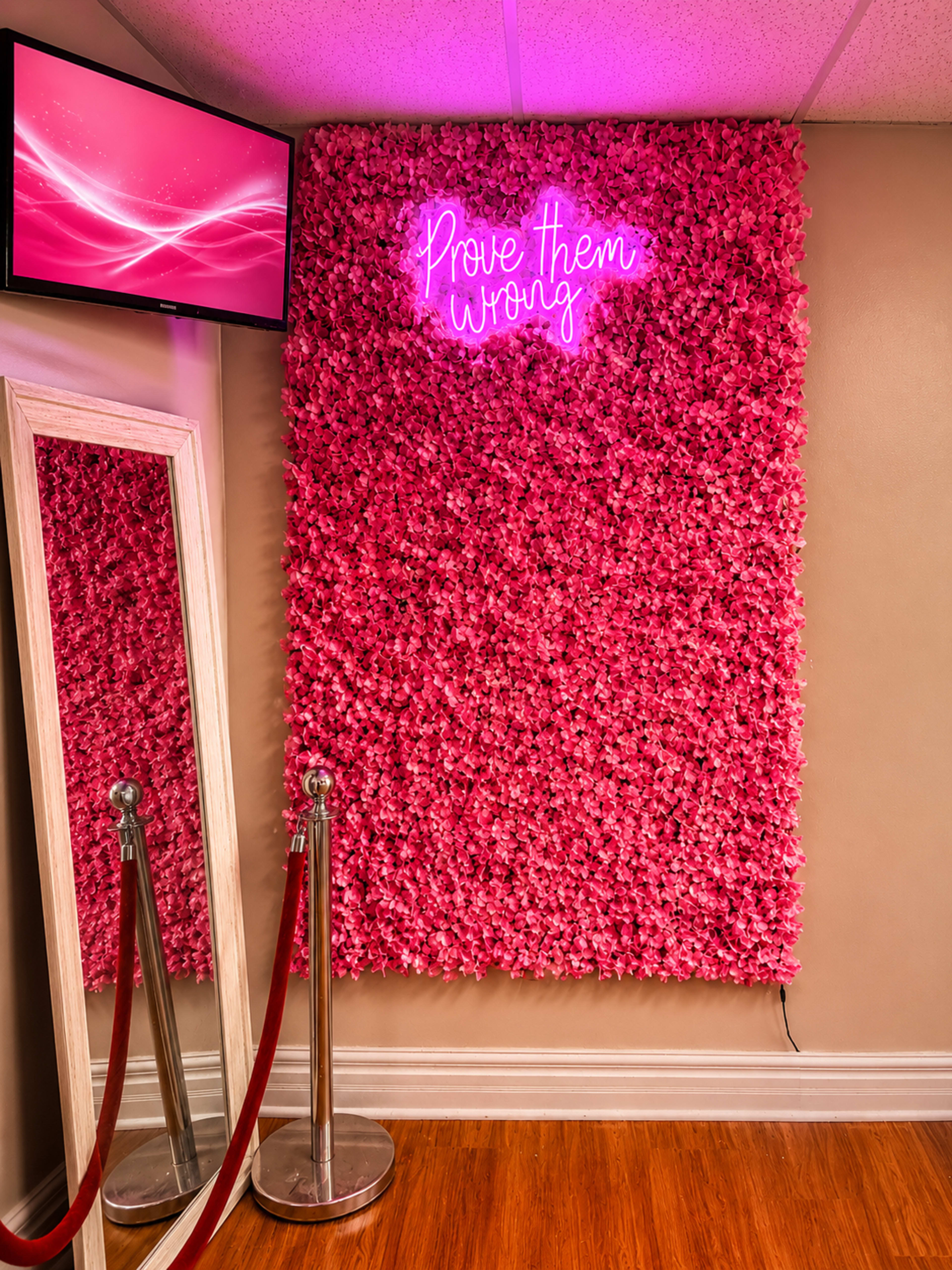 The image shows a pink floral wall with a neon sign that reads "Prove them wrong," alongside a mirror and a television screen in a well-lit interior space.