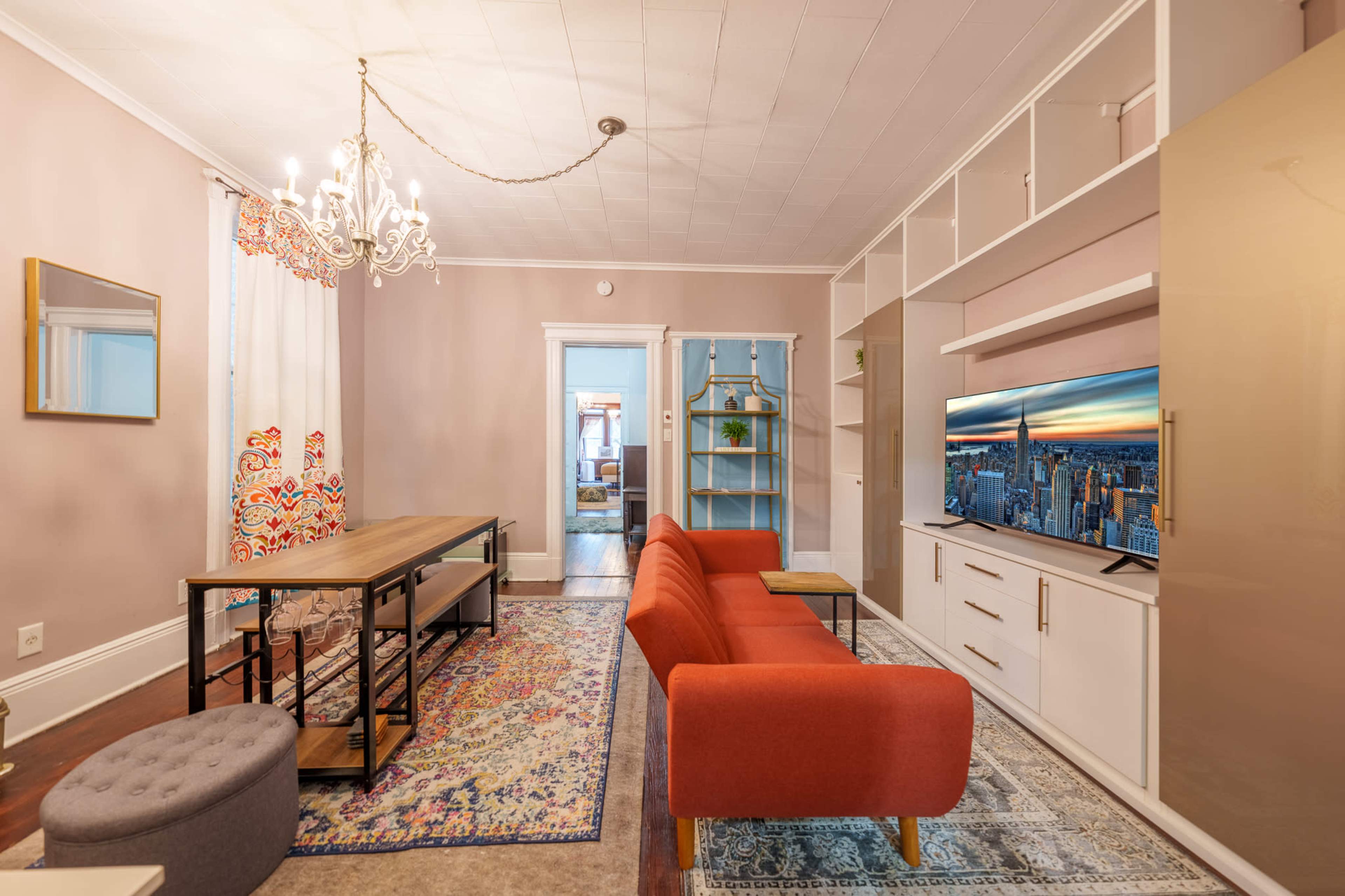 The image depicts a living room with a red sofa, a dining table, a television stand, and decorative elements including a chandelier and patterned curtains.