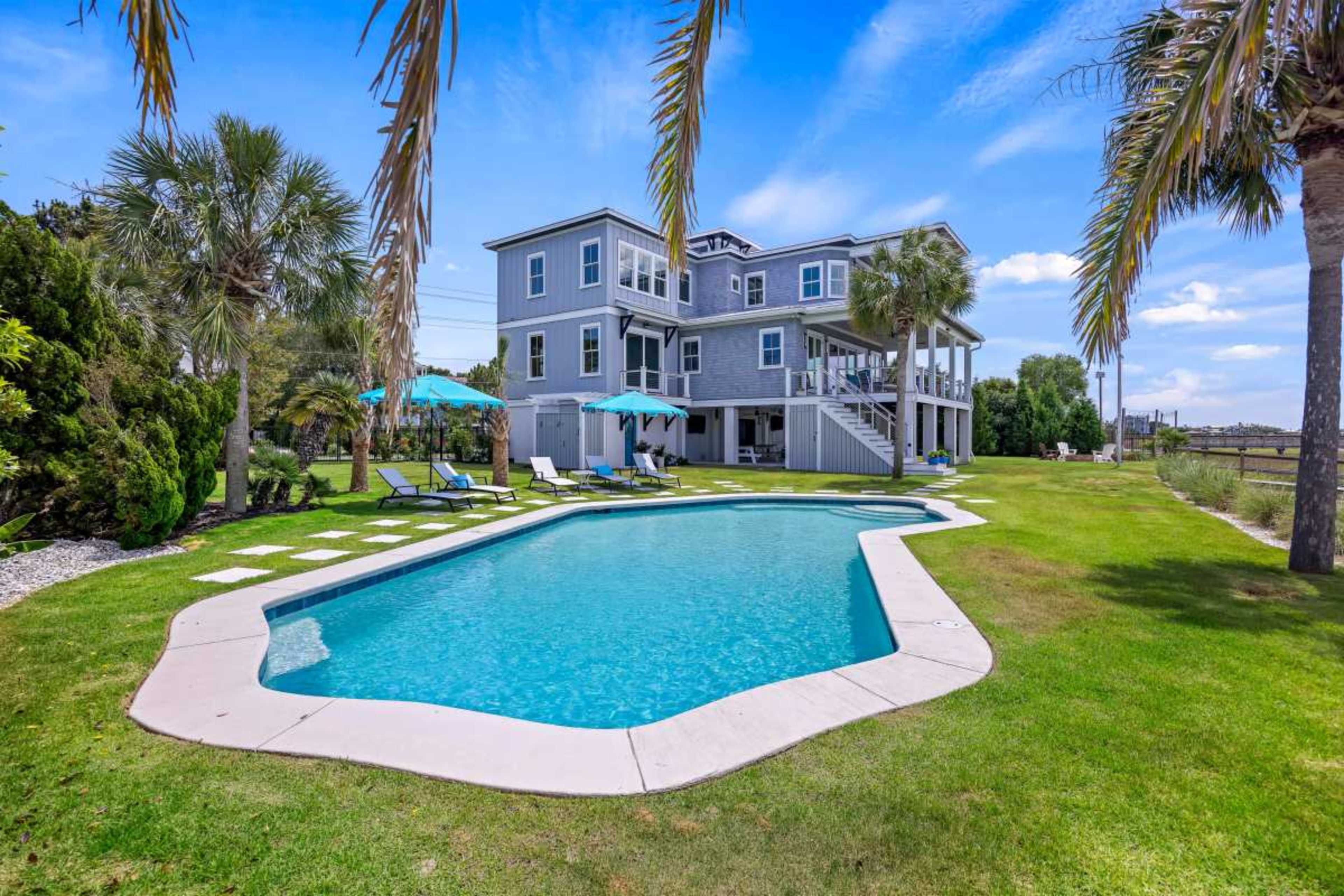 A two-story gray house with a swimming pool and palm trees is situated on a grassy area, surrounded by lounge chairs and umbrellas.