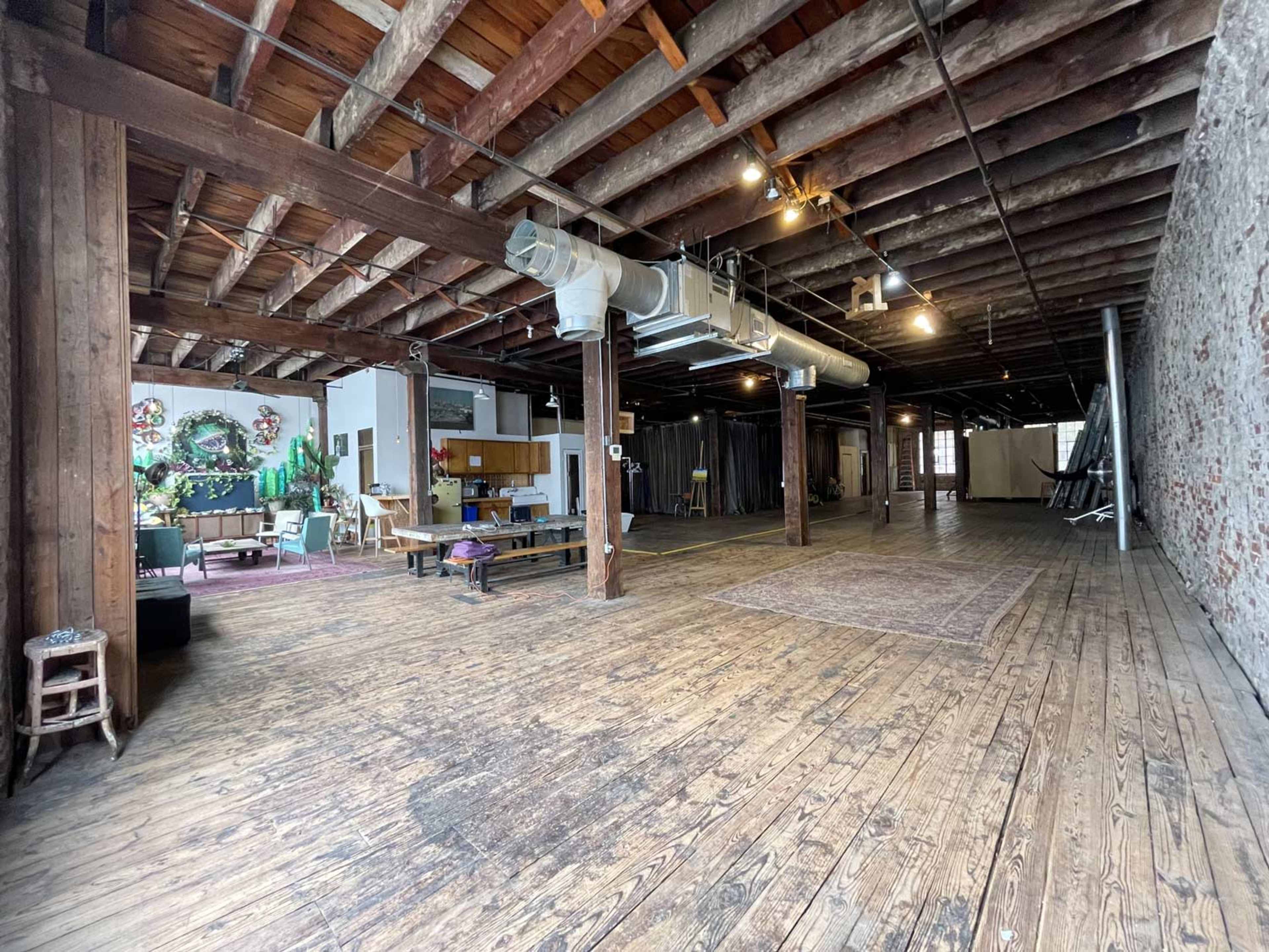 The image shows a spacious, industrial-style loft with exposed wooden beams and a wooden floor, featuring minimal furniture and murals on the walls.