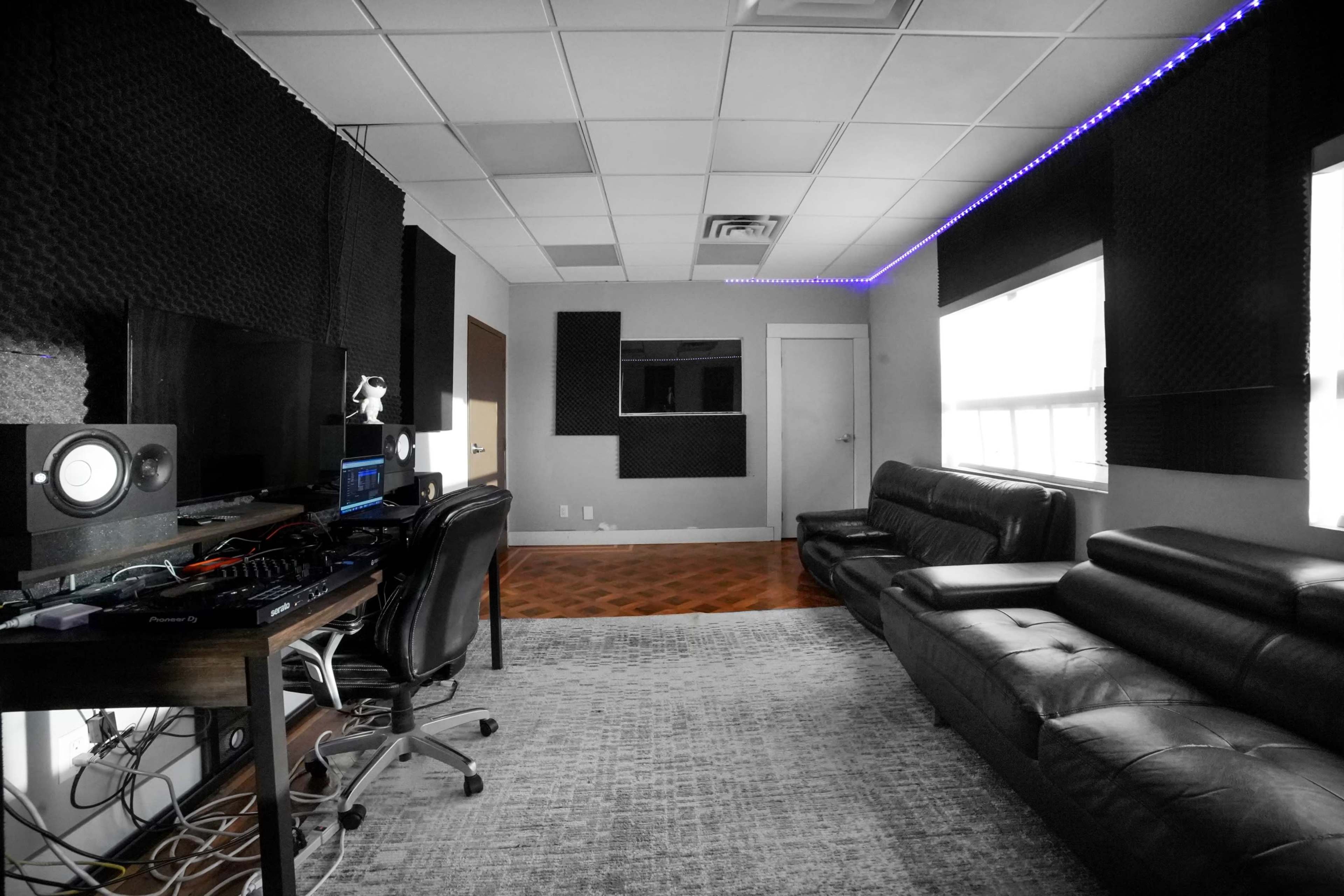 A music production studio featuring a desk with audio equipment, a black leather couch, and soundproofing panels on the walls.
