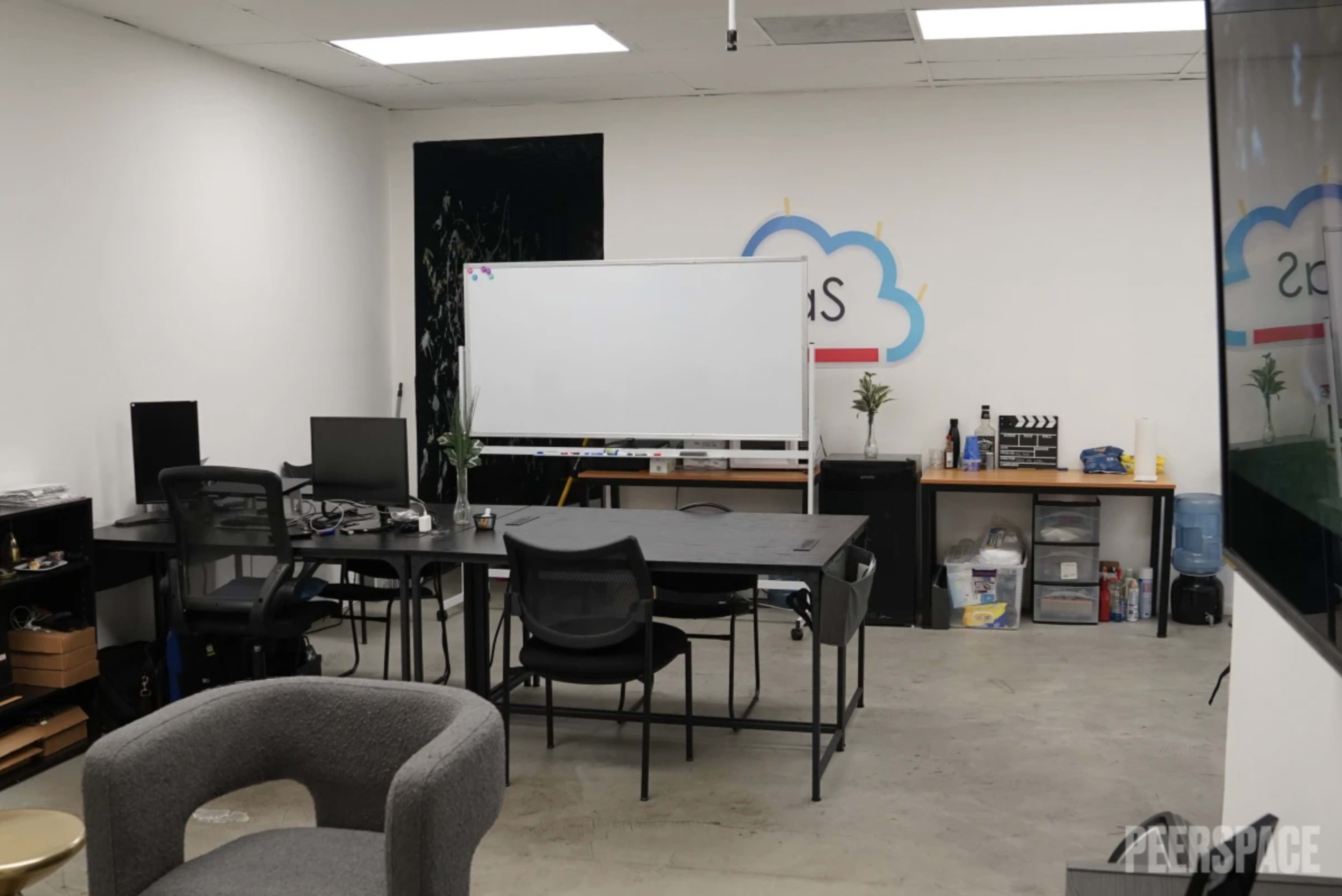 A workspace with a large table, several chairs, a whiteboard, and shelving units containing various items.