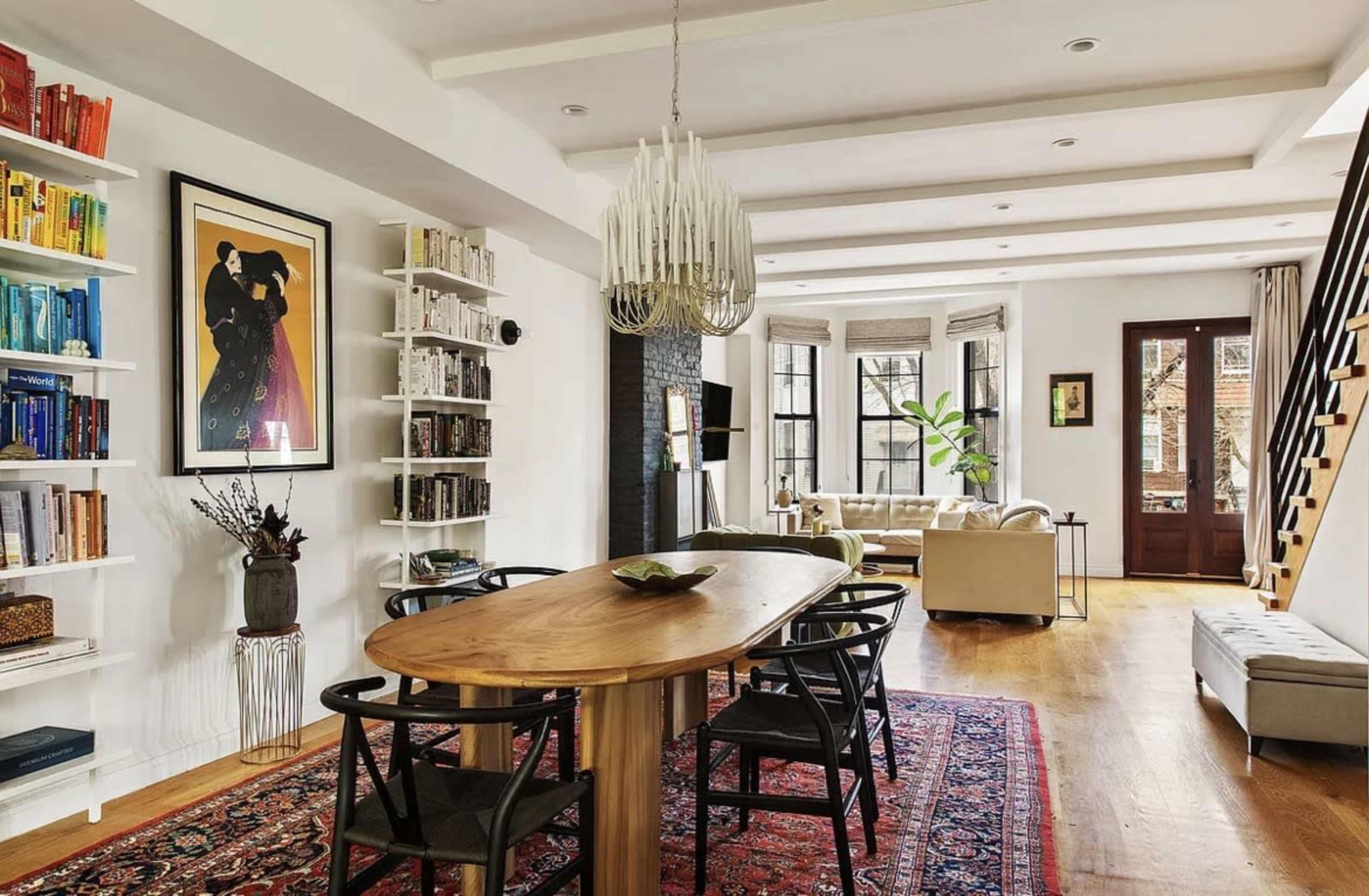 The image shows a spacious, modern living and dining area with a large wooden dining table, bookshelves filled with books, and a cozy seating arrangement.