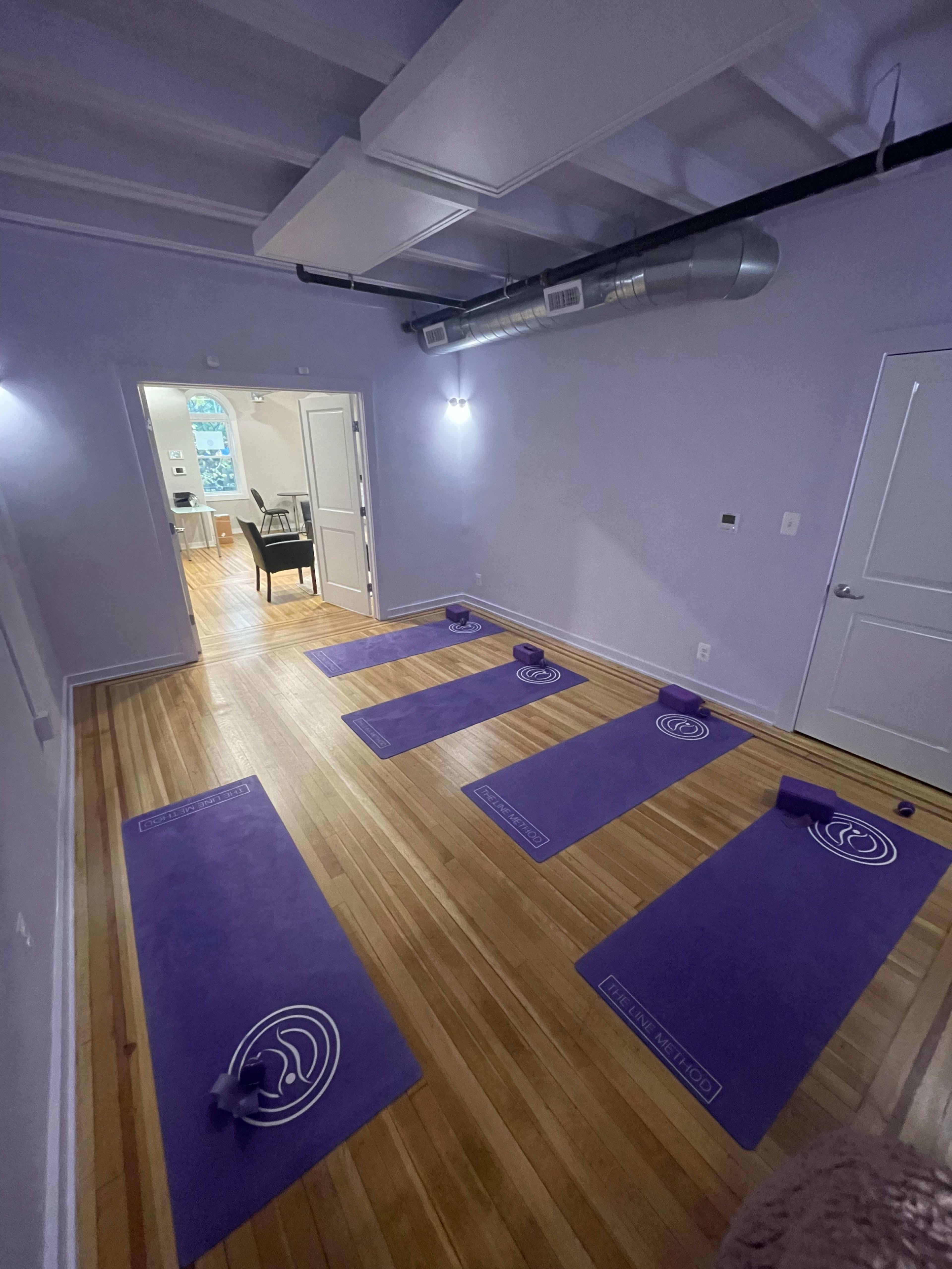 The image shows a light purple yoga studio with six yoga mats arranged on the wooden floor.