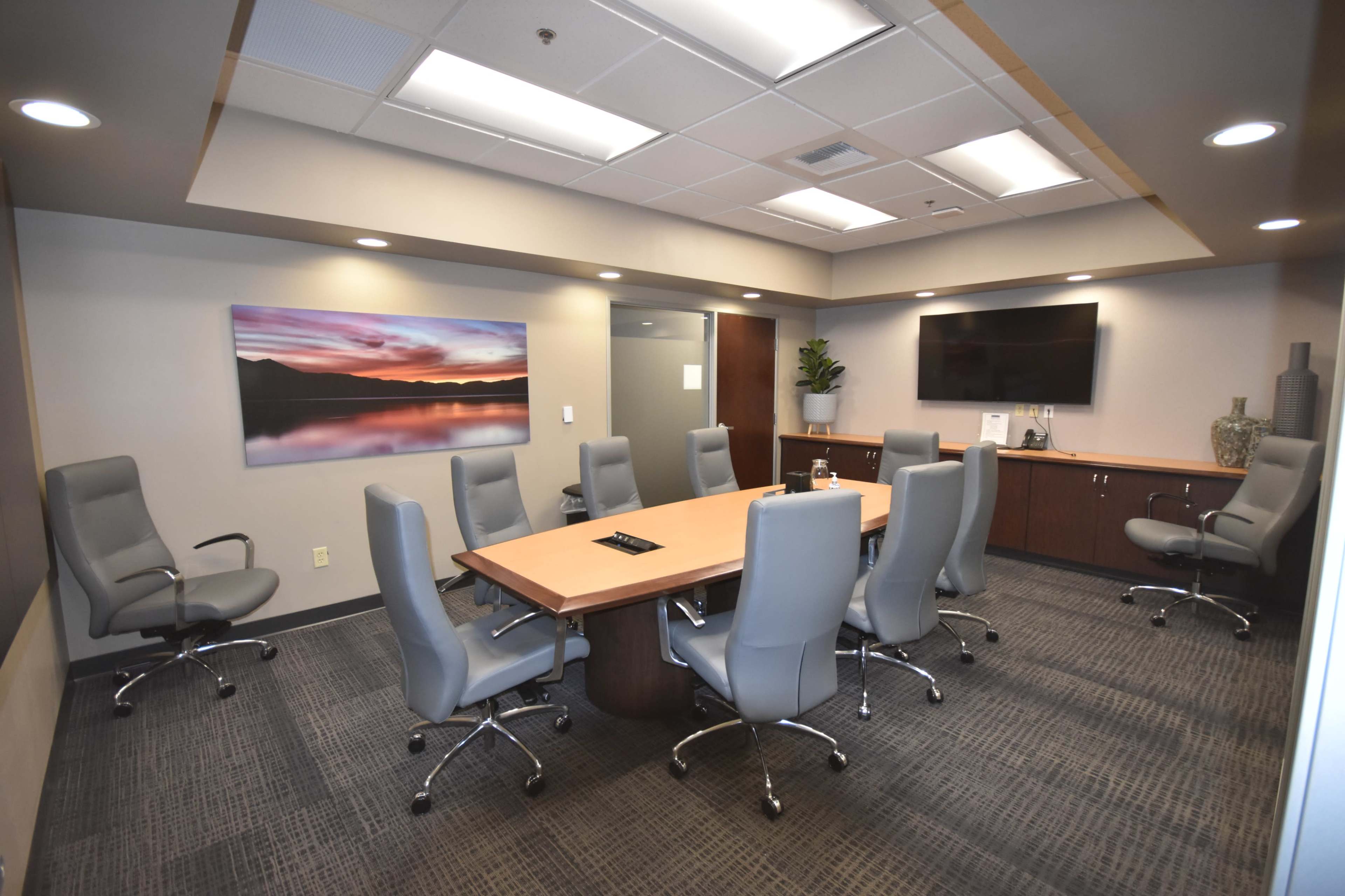 The image shows a modern conference room equipped with a large wooden table surrounded by ten gray swivel chairs, along with a wall-mounted television and a landscape painting.