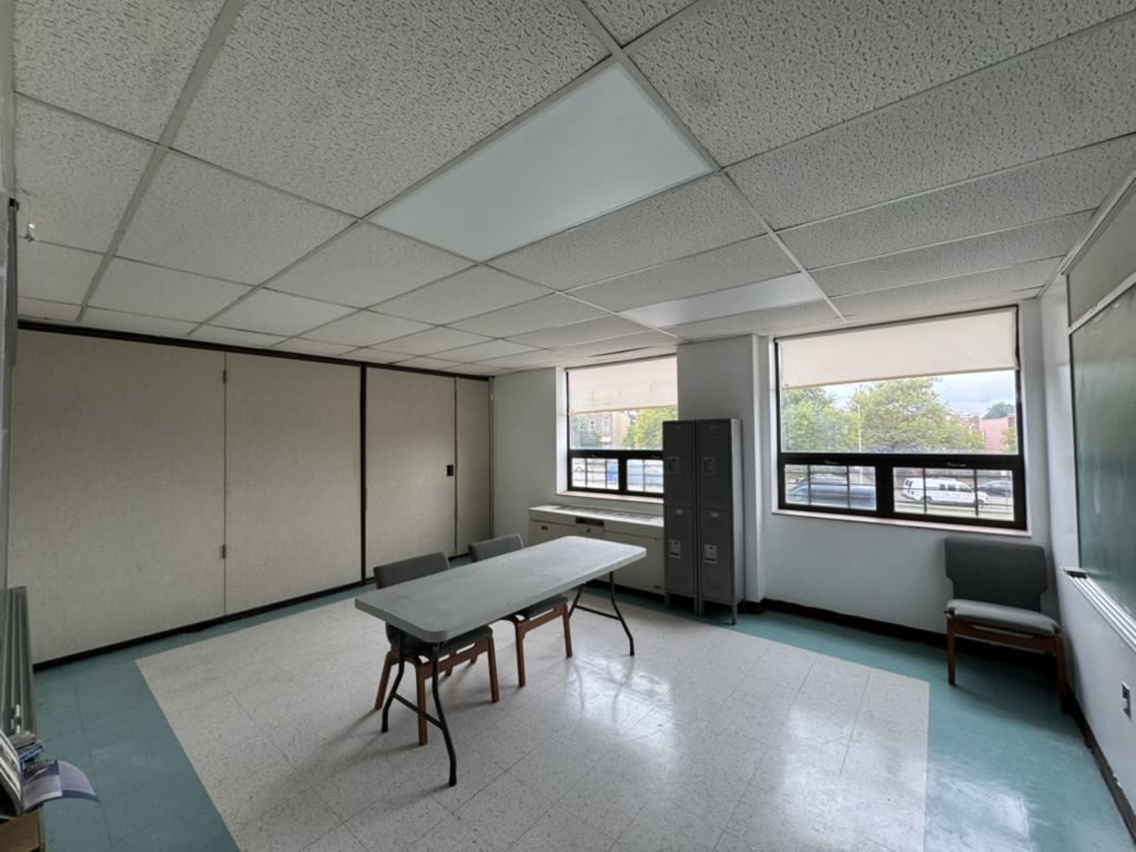 An empty classroom features a central table surrounded by chairs, with windows on either side and a green chalkboard at the back.
