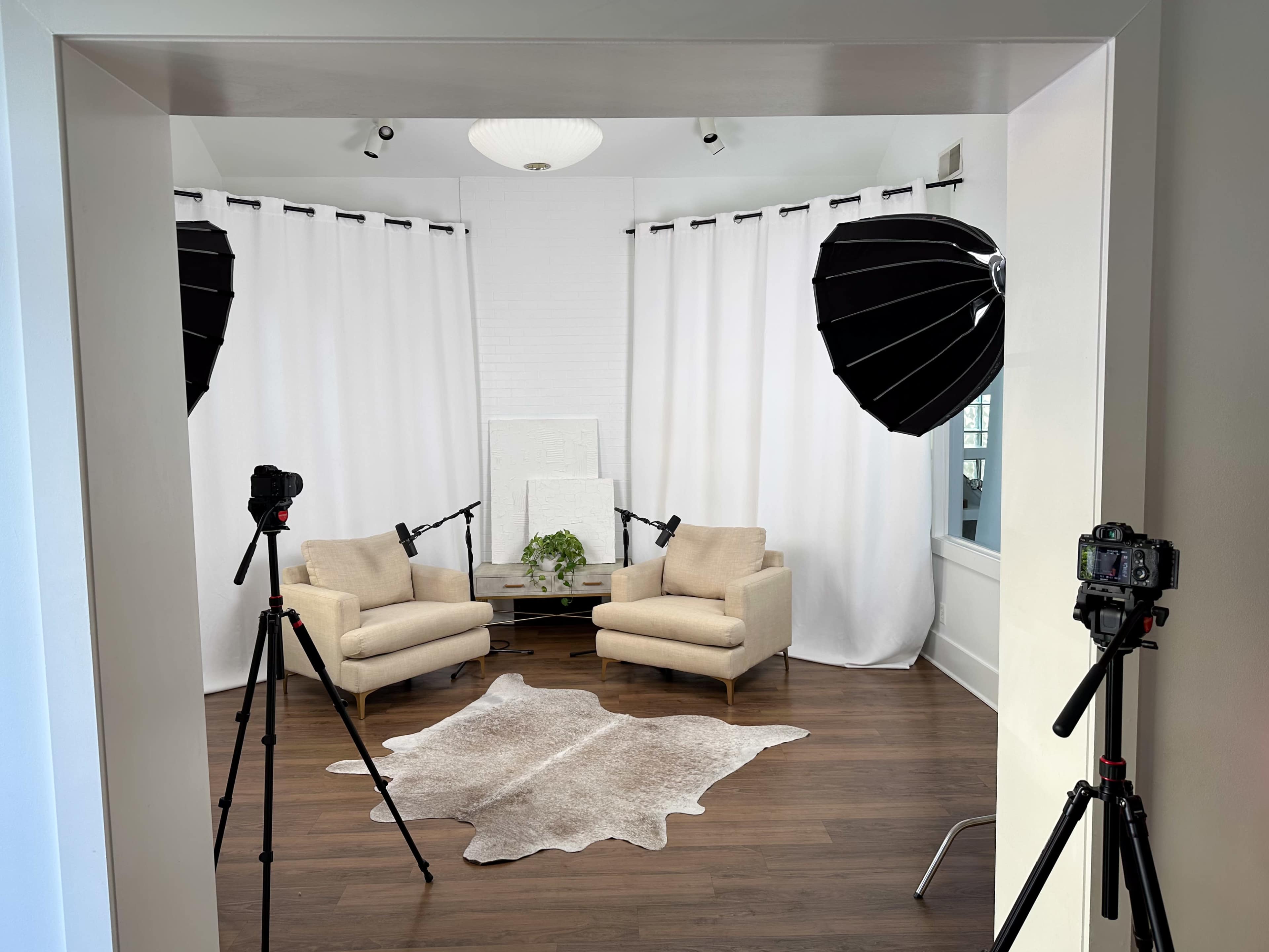 A well-lit studio setup features two beige chairs, a coffee table, and soft lighting equipment arranged in front of white curtains.