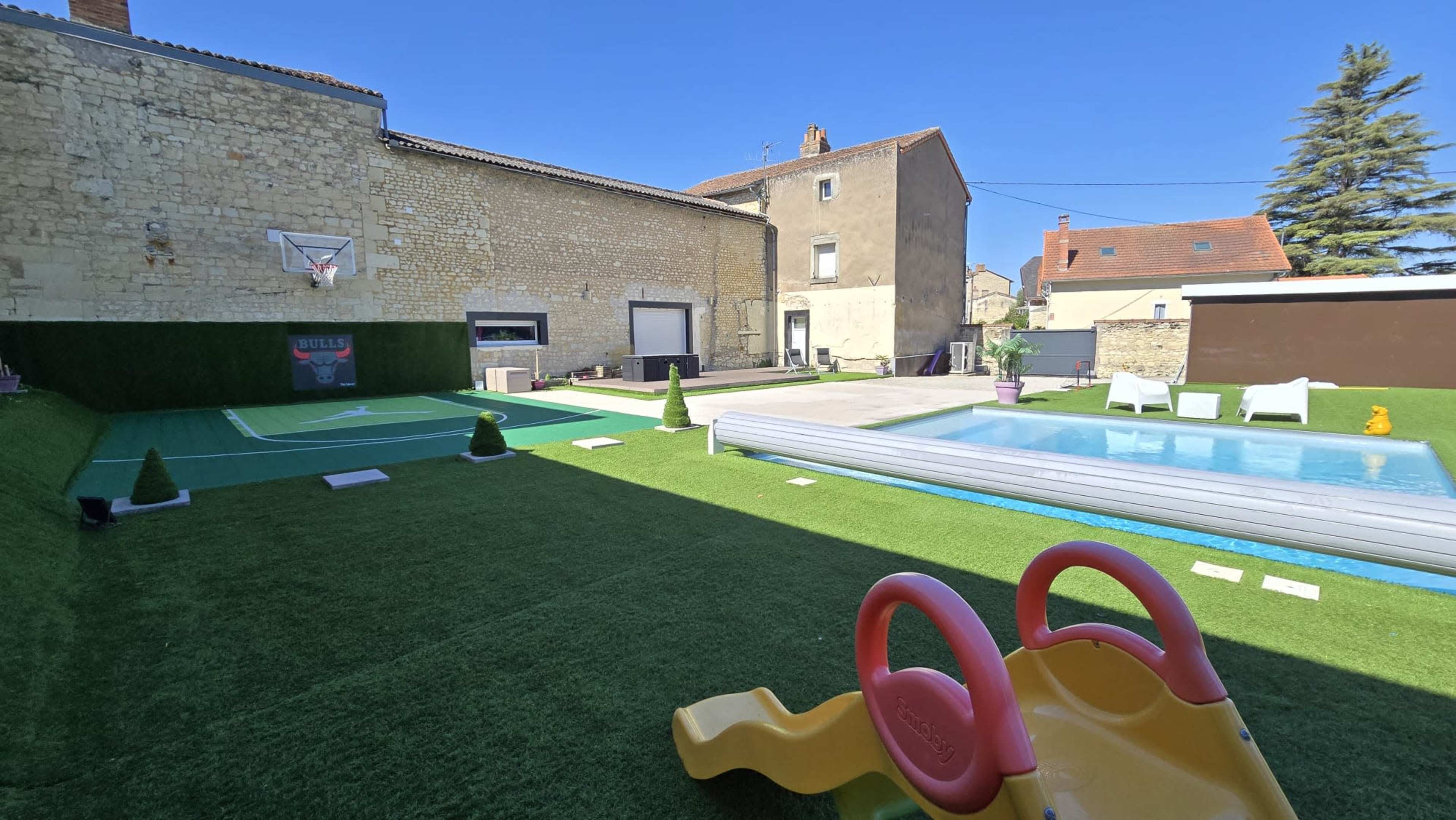 The image shows a spacious outdoor area featuring a green turf lawn, a basketball hoop, a swimming pool, and a playground slide, surrounded by residential buildings.