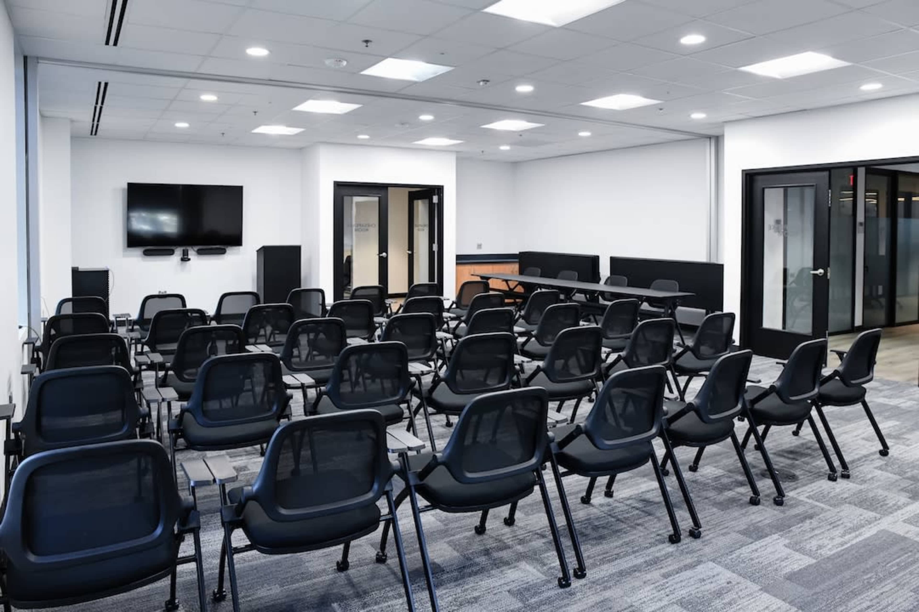 The image shows a modern meeting room set up with rows of black chairs facing a large screen and a conference table.