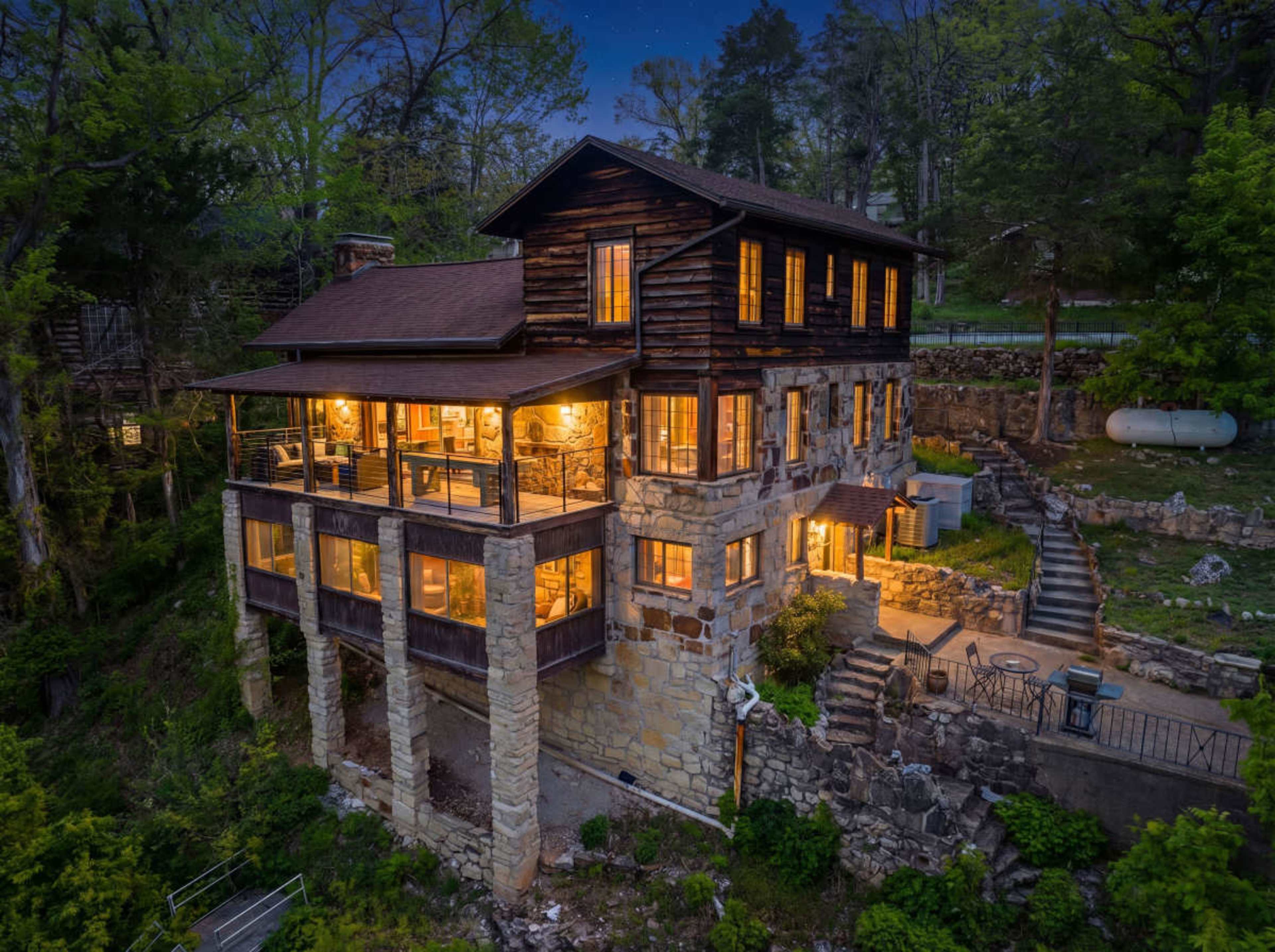 The image shows a multi-level house made of wood and stone, elevated on stilts, with illuminated windows surrounded by greenery at dusk.