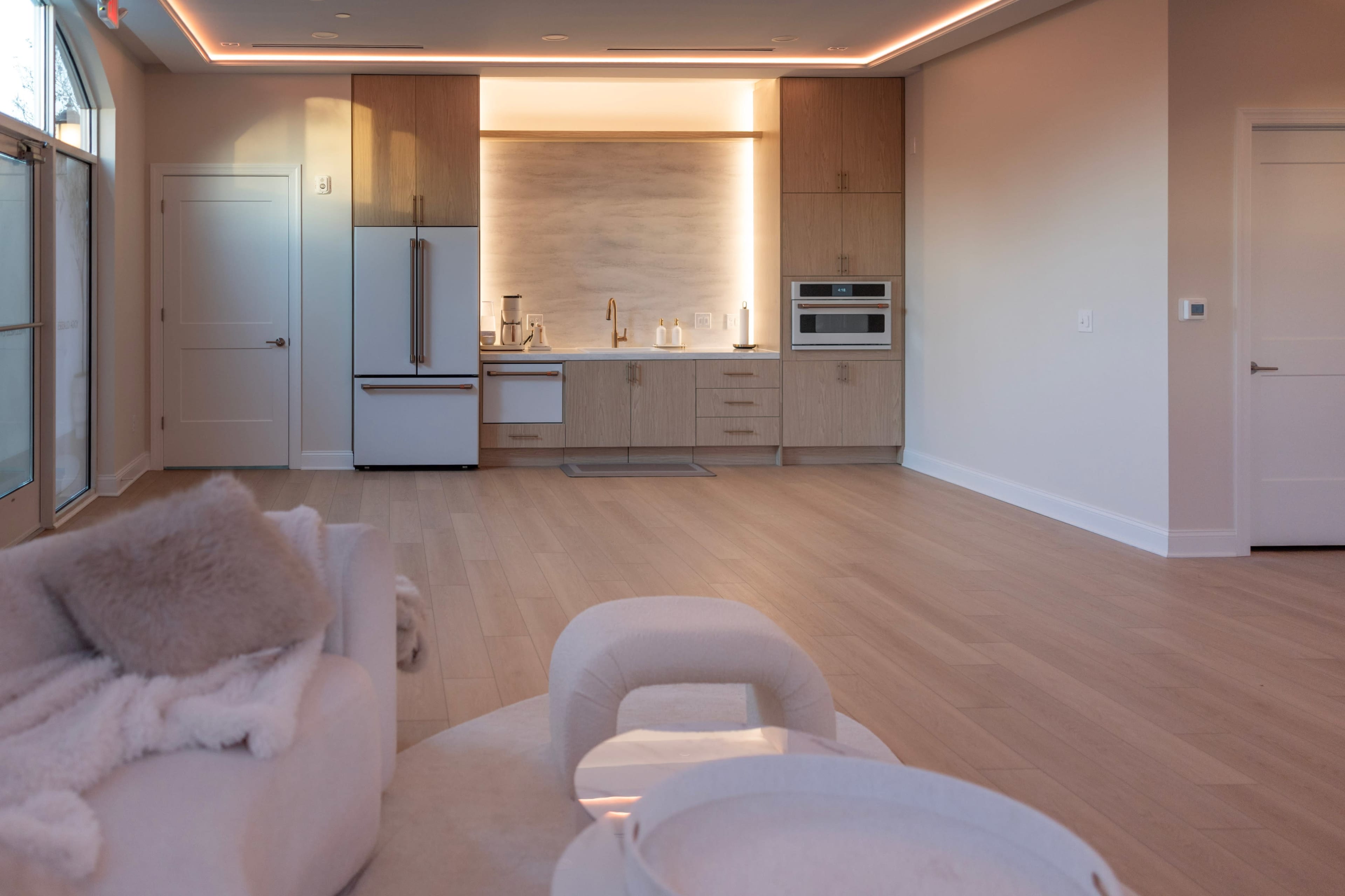 The image shows a modern, open-concept living space featuring a sleek kitchen with wood cabinetry, stainless steel appliances, and a cozy seating area with a light-colored sofa.