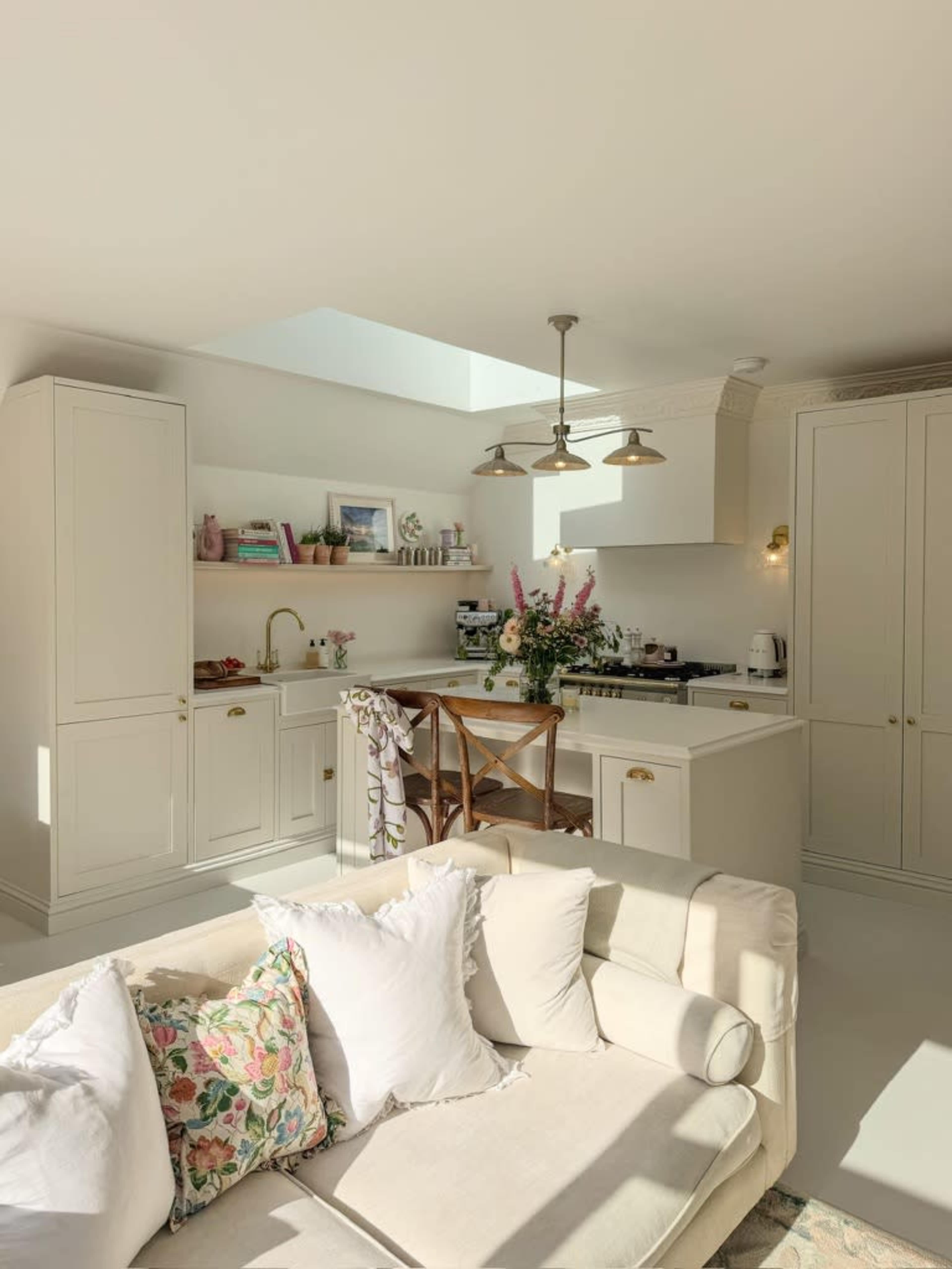 A bright kitchen features light-colored cabinets, a central island with flowers, and sunlight streaming through a skylight, with a cozy sofa nearby.
