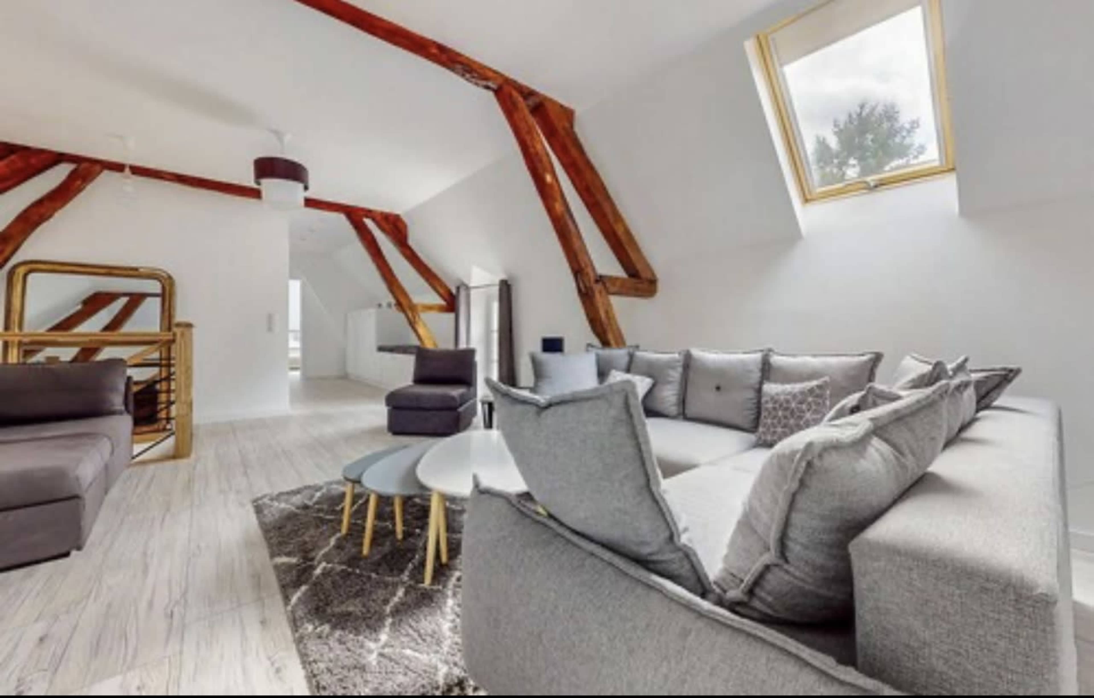 The image shows a modern, airy living room with vaulted ceilings, wooden beams, a sectional sofa, and a set of coffee tables on a textured rug.