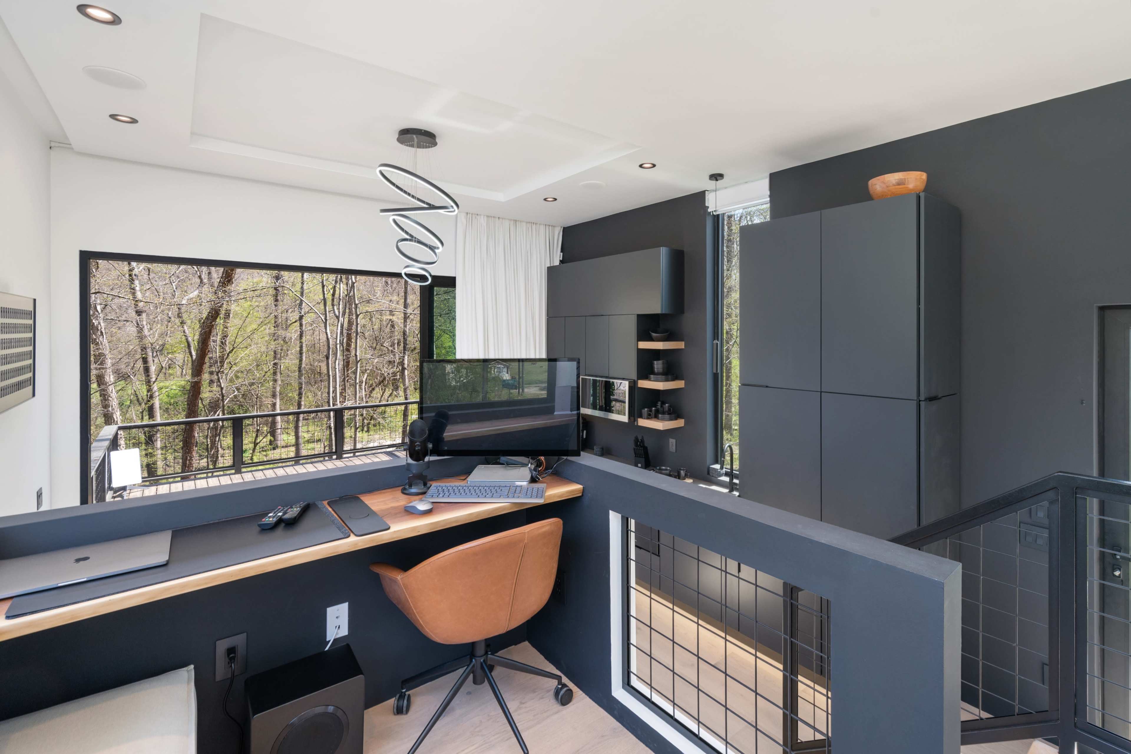 A modern workspace features a desk with a computer, shelves, and a view of trees through large windows.