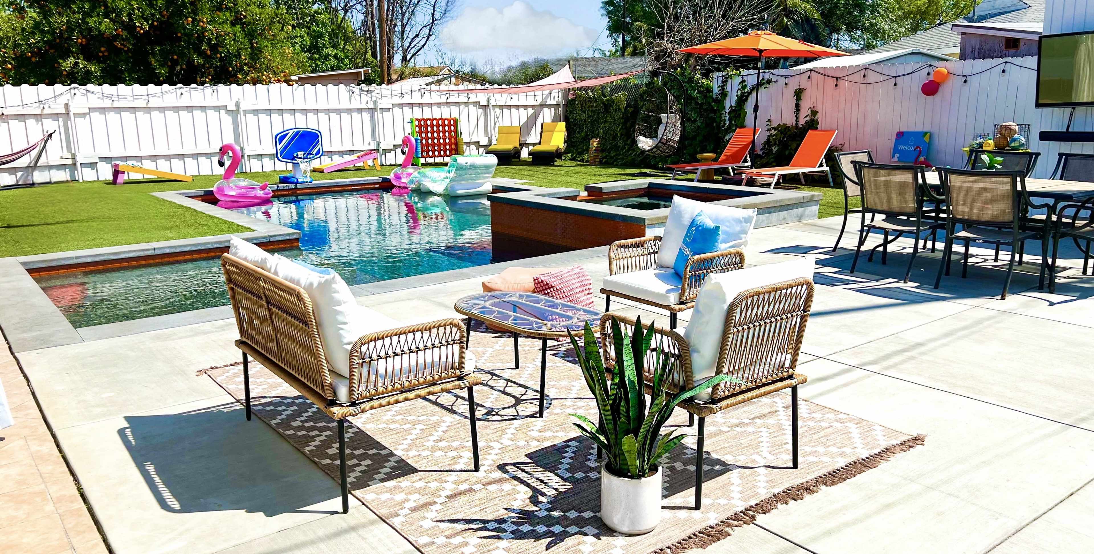 The image shows a backyard scene featuring a swimming pool surrounded by lounge chairs, a fire pit, and inflatable pool toys, with a colorful umbrella providing shade.