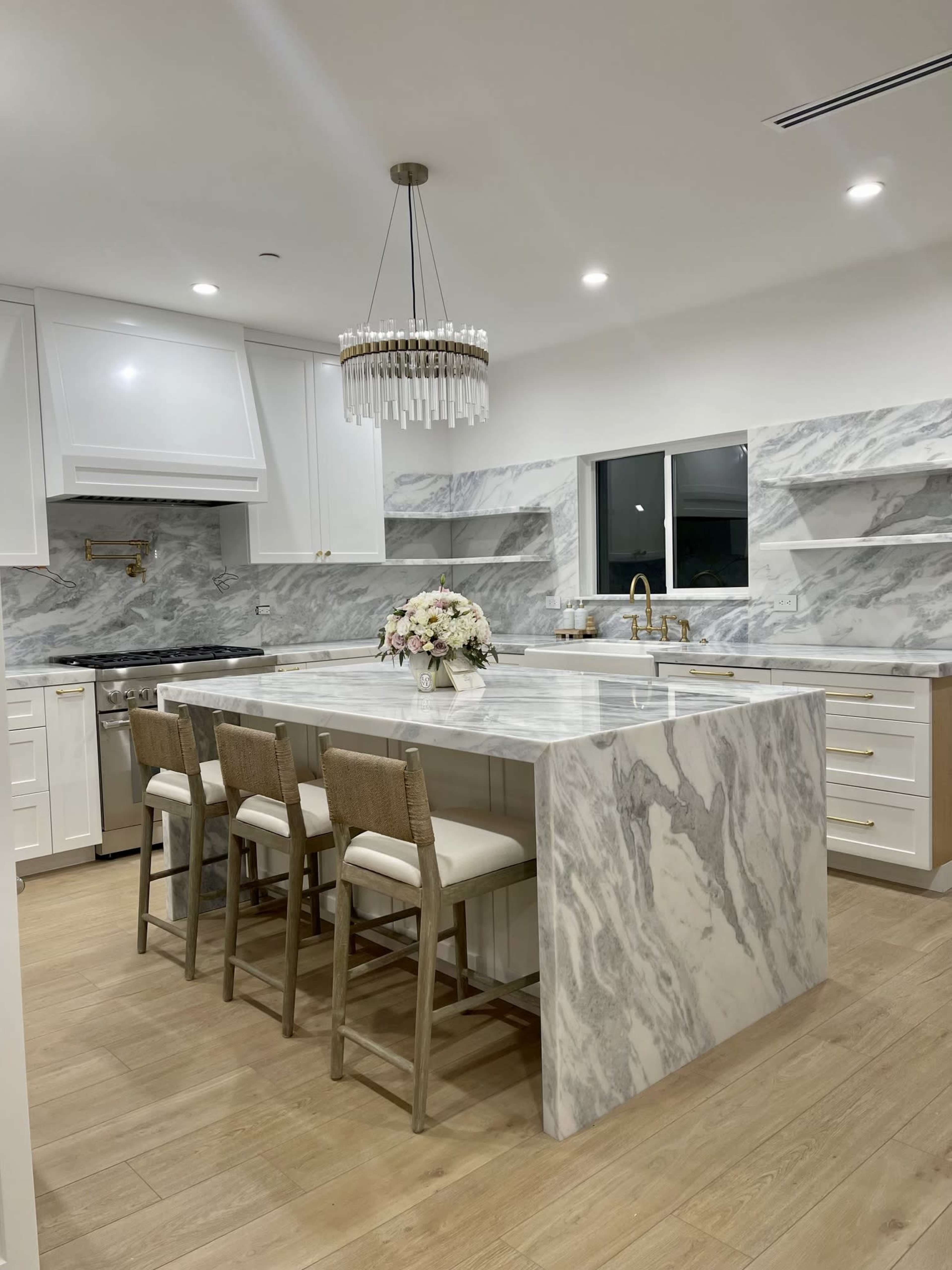A modern kitchen features a large marble island with seating, white cabinetry, and a chandelier above.