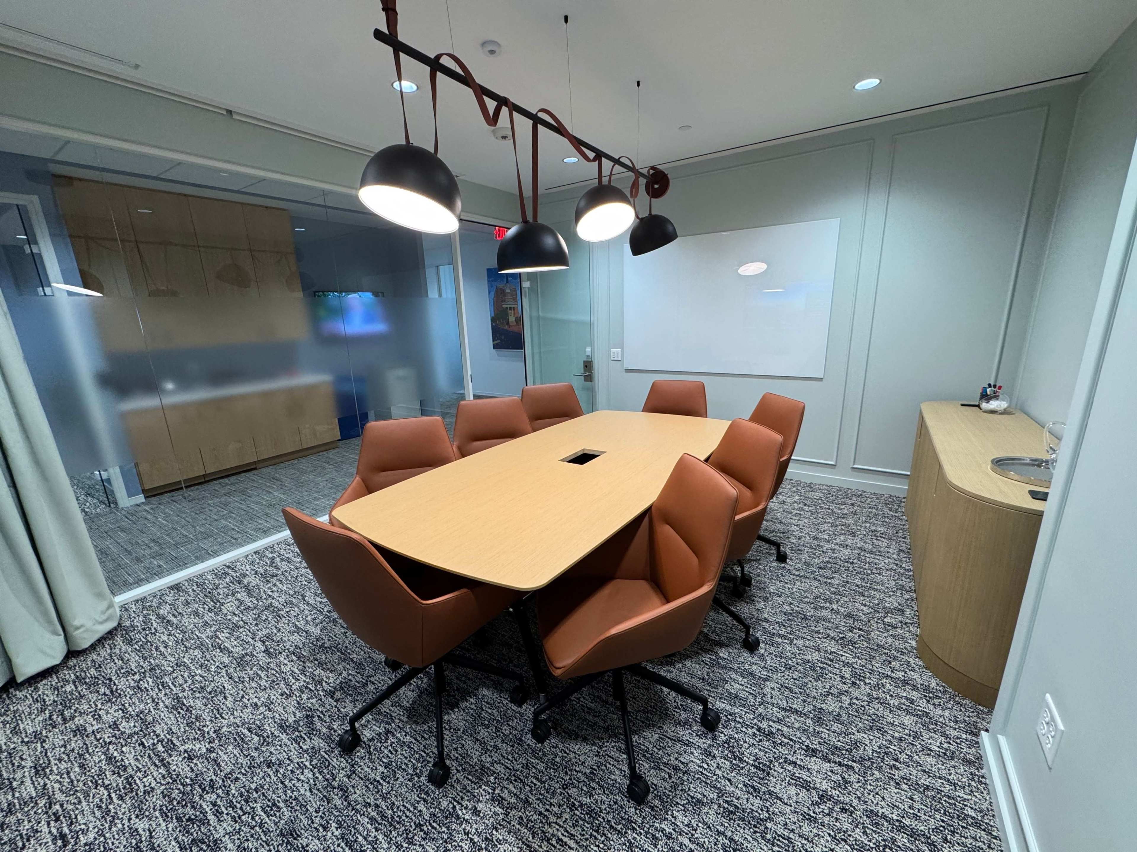 A modern conference room features a large table surrounded by six brown chairs, with pendant lighting and a glass wall on one side.