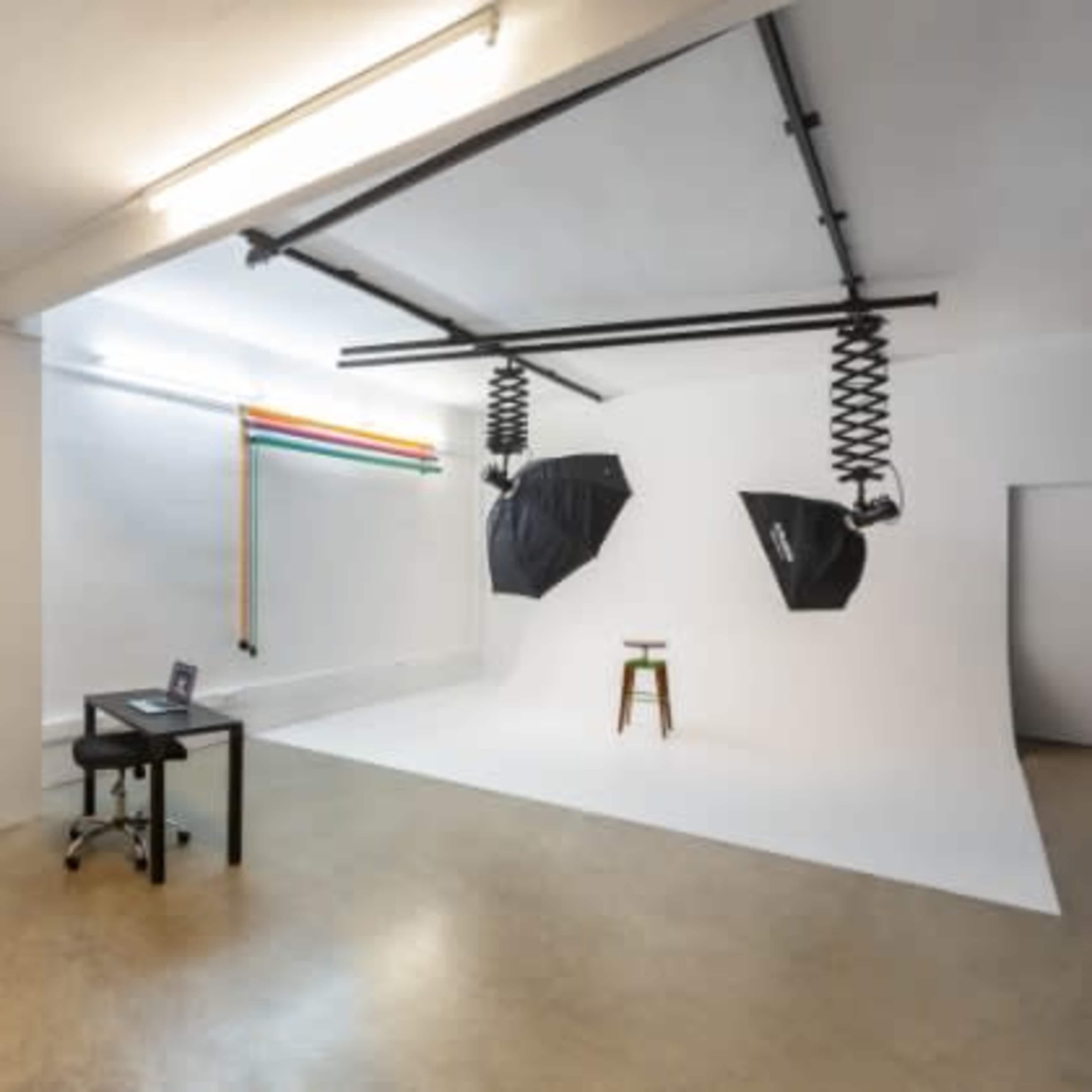 The image shows a minimalist photography studio featuring a white backdrop, two overhead lights, a stool, and a desk with a computer.