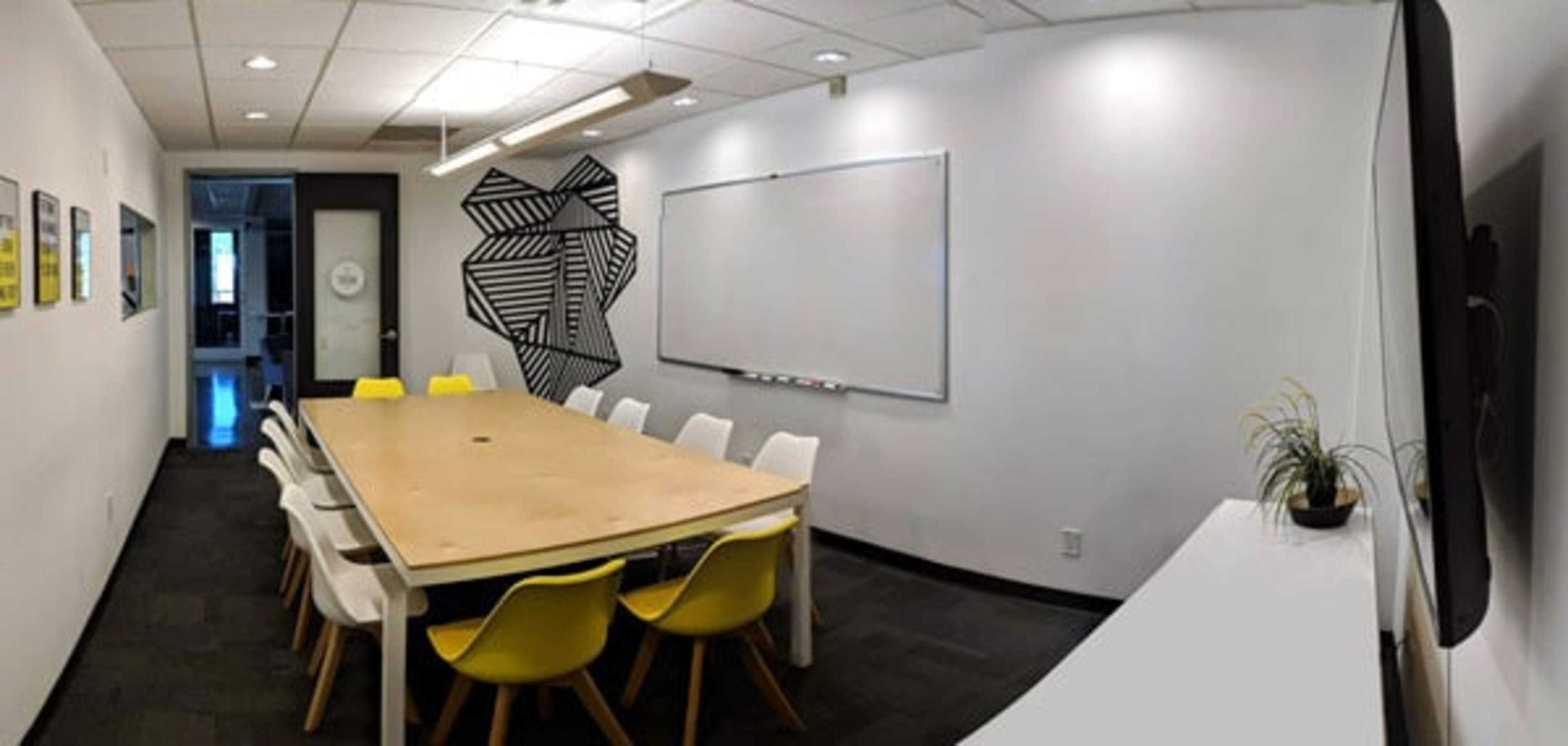 A modern conference room with a large wooden table surrounded by white and yellow chairs, a whiteboard on one wall, and a minimalist plant on a side shelf.