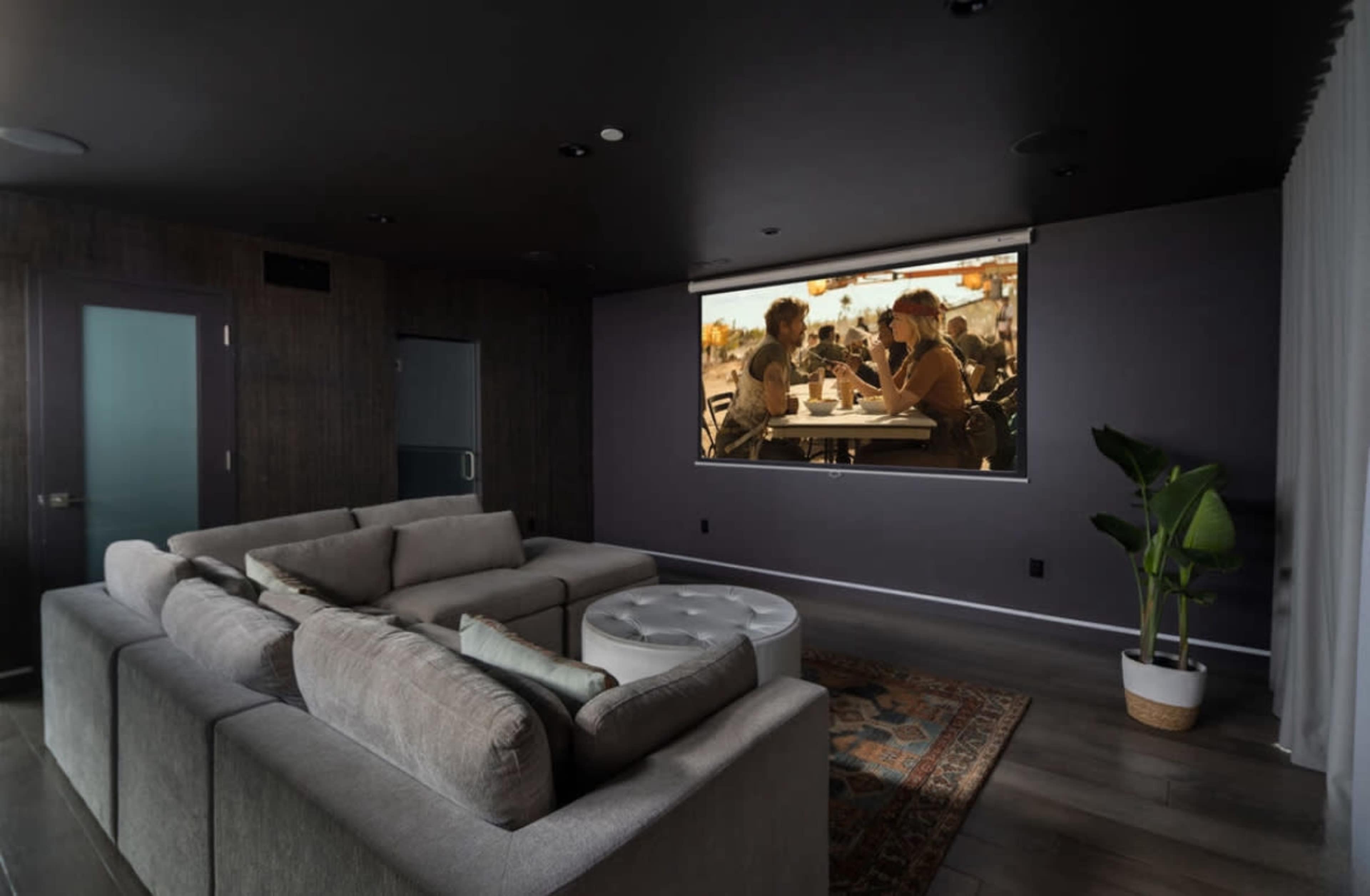 A modern living room features a large sectional sofa facing a projection screen displaying two people at a café.