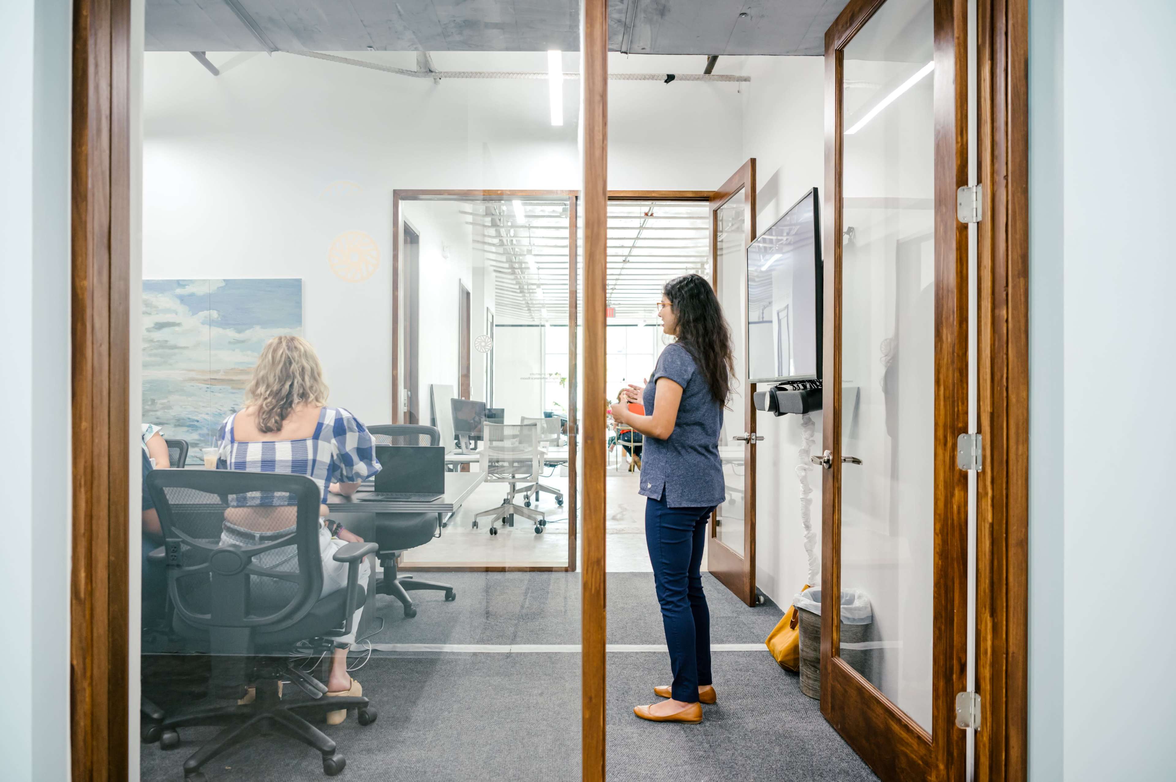 A woman stands in a glass-walled office, speaking to a group of colleagues seated at a conference table.