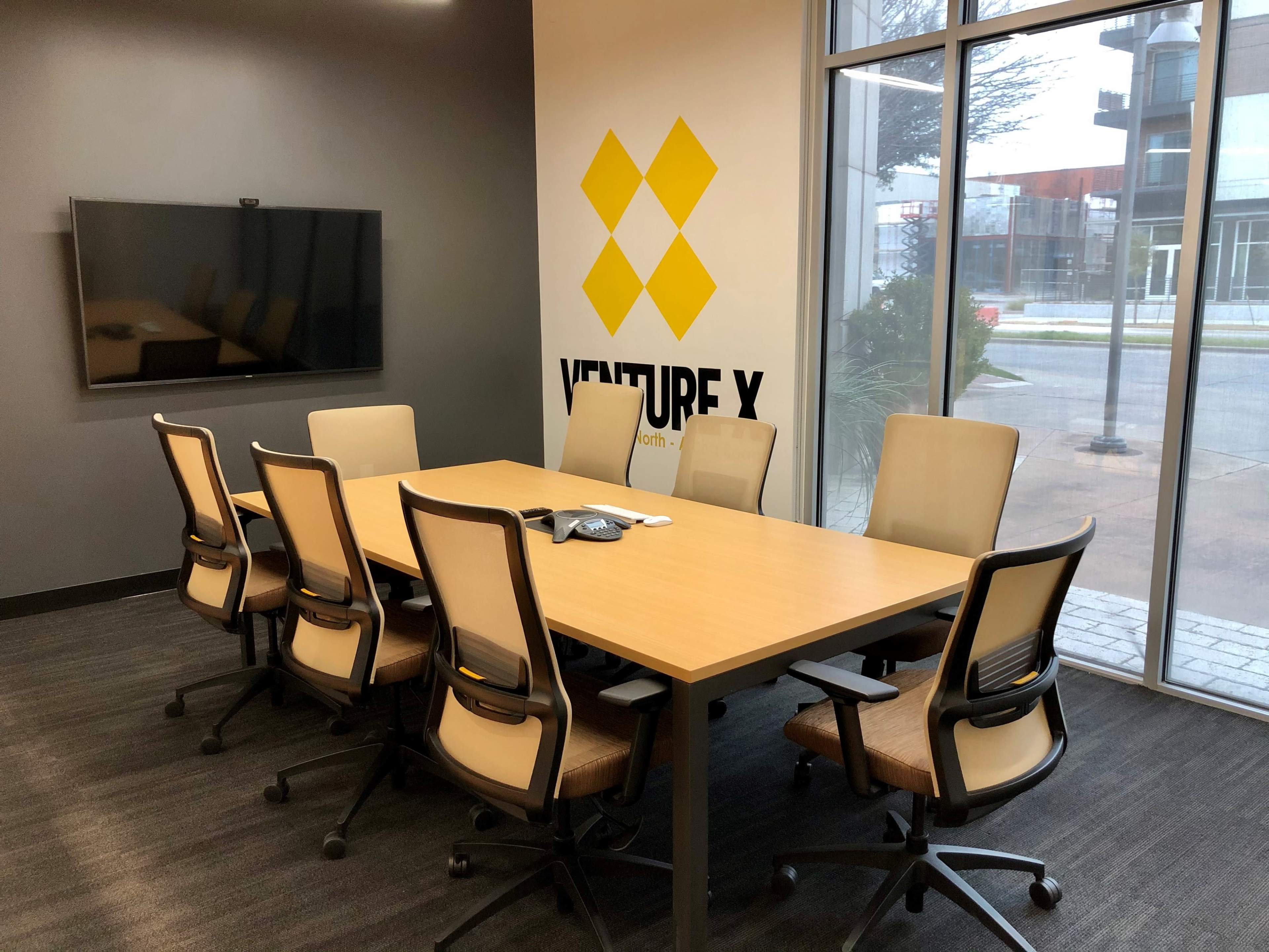 A conference room features a long wooden table surrounded by eight chairs, with a large monitor on the wall and a company logo displayed prominently.