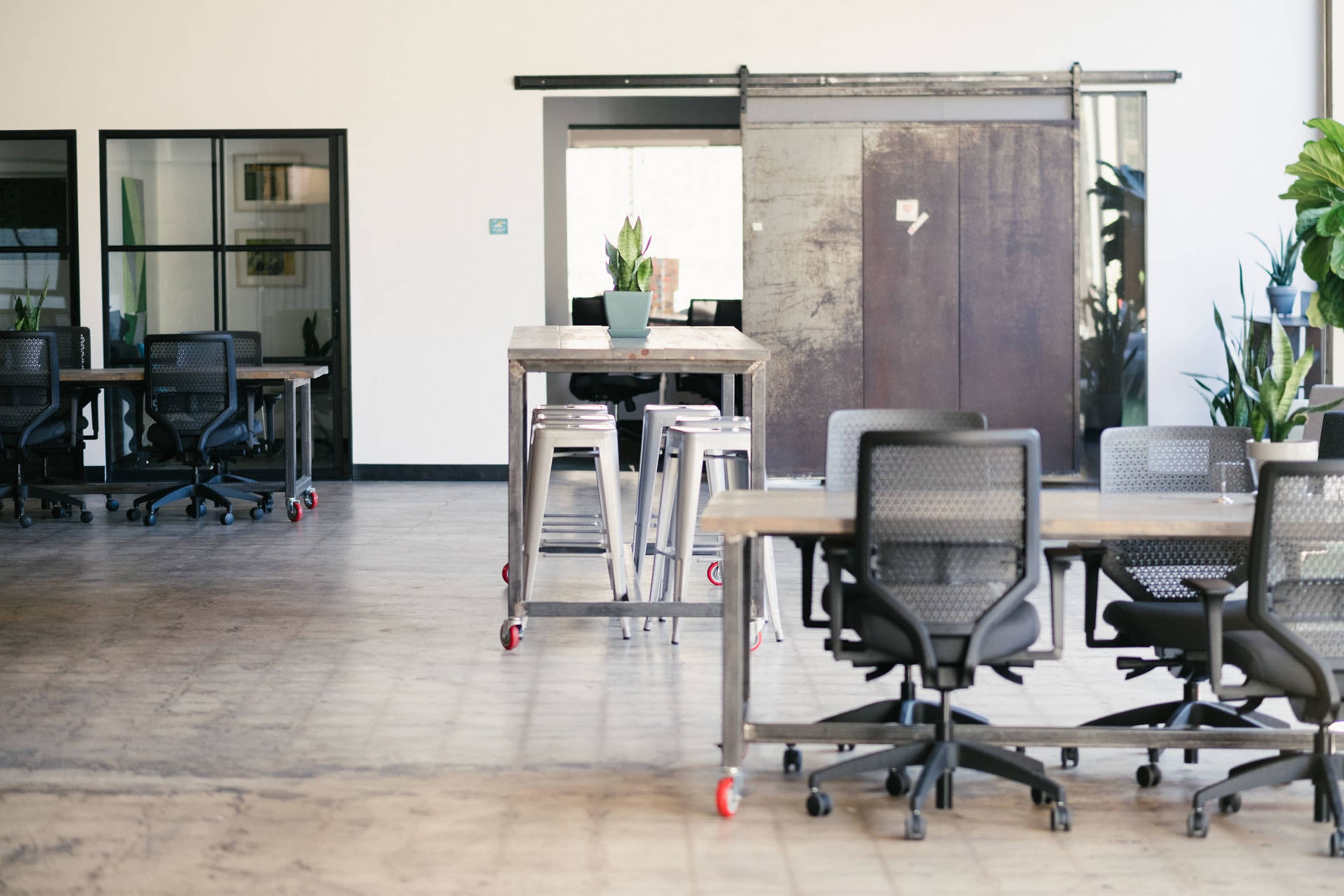 The image shows a spacious office interior with several desks, rolling chairs, and a large sliding door, complemented by potted plants.