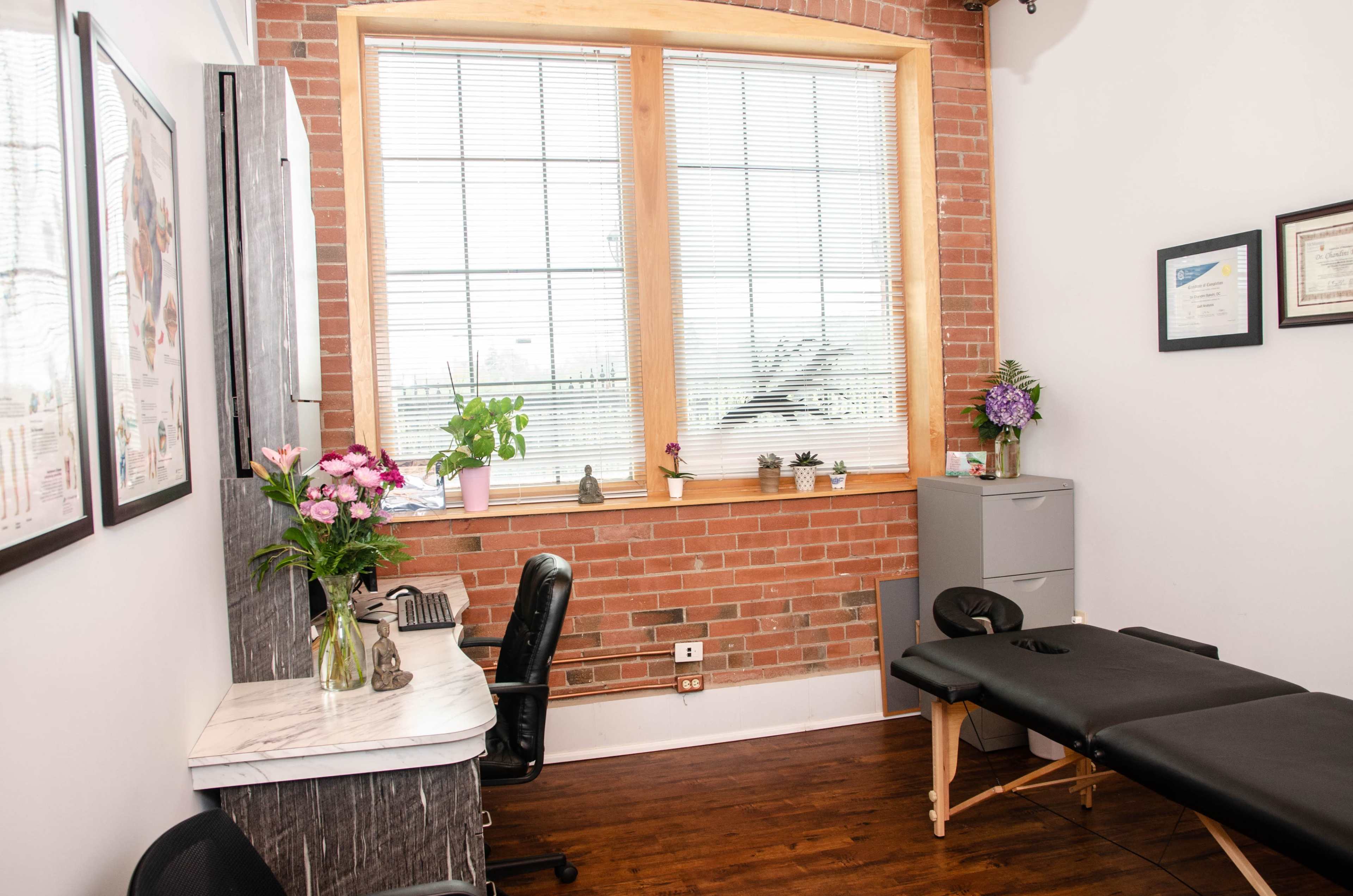 A treatment room featuring a black massage table, a small desk with flowers, and a large window with blinds.
