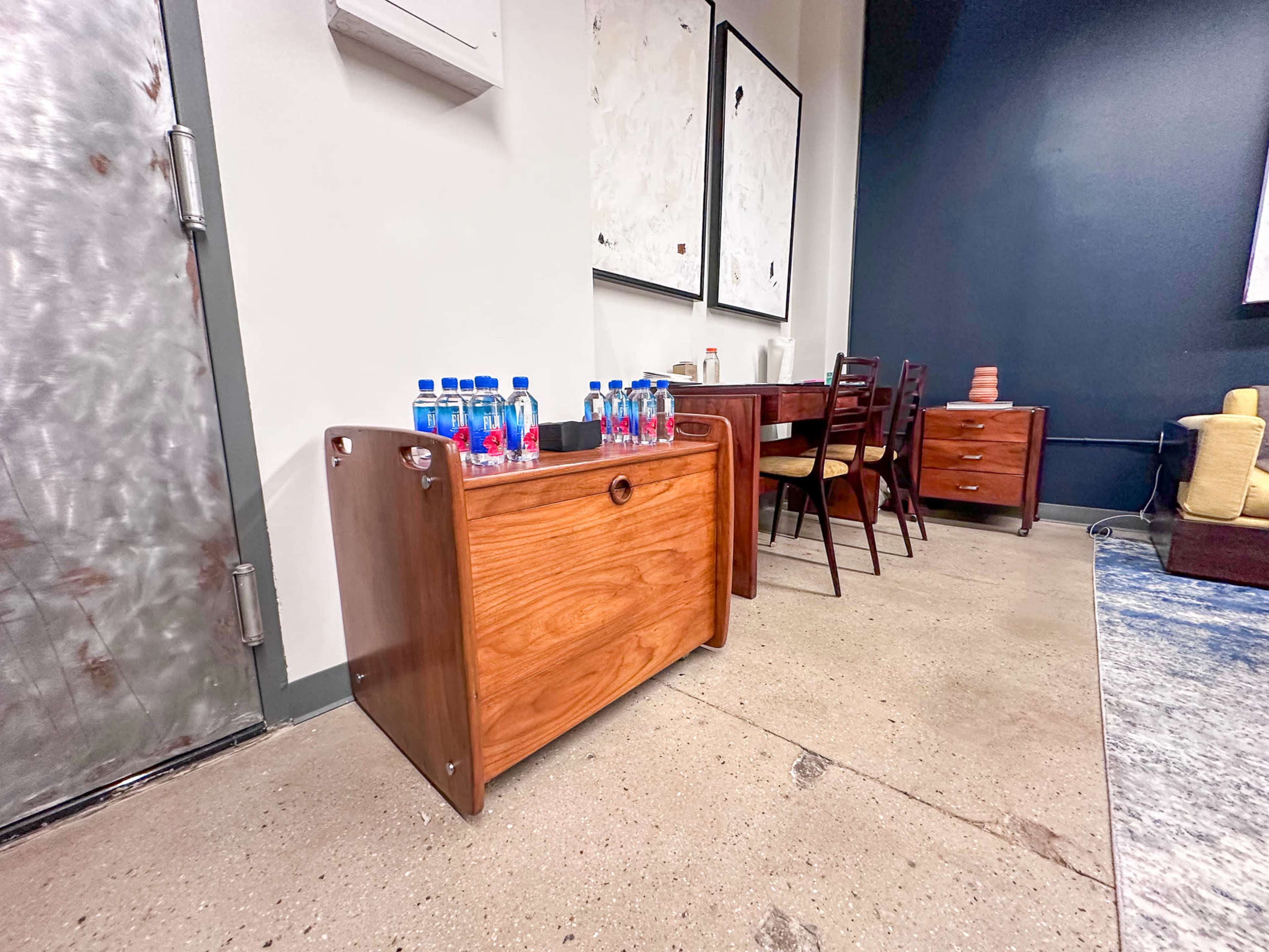 The image shows a wooden storage chest with a row of bottled water placed on top, located next to a dining table and chairs in a room with a gray wall.