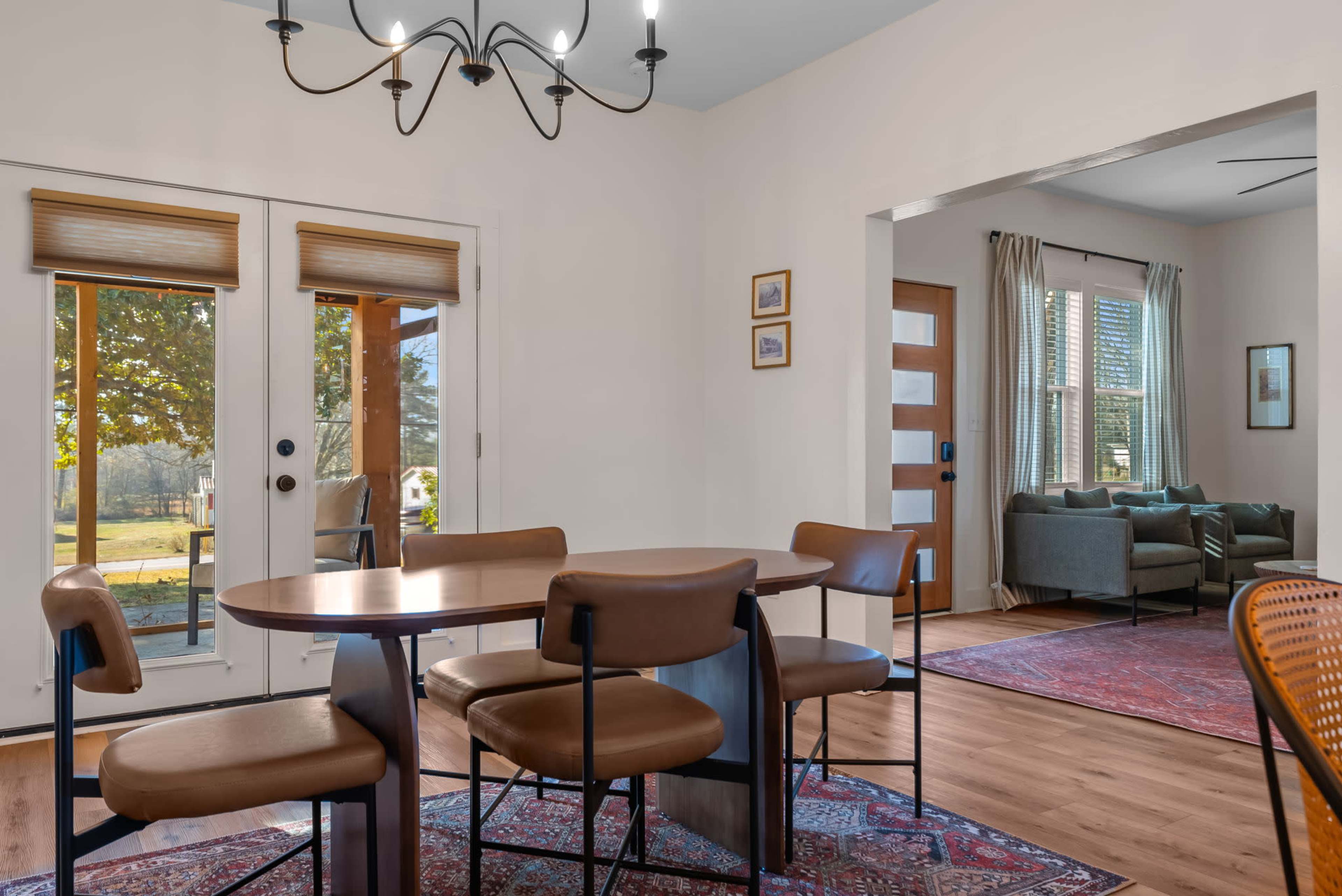 A dining area with a round table and chairs is visible, featuring large windows that open to an outdoor view and a doorway leading to a living space with a couch.