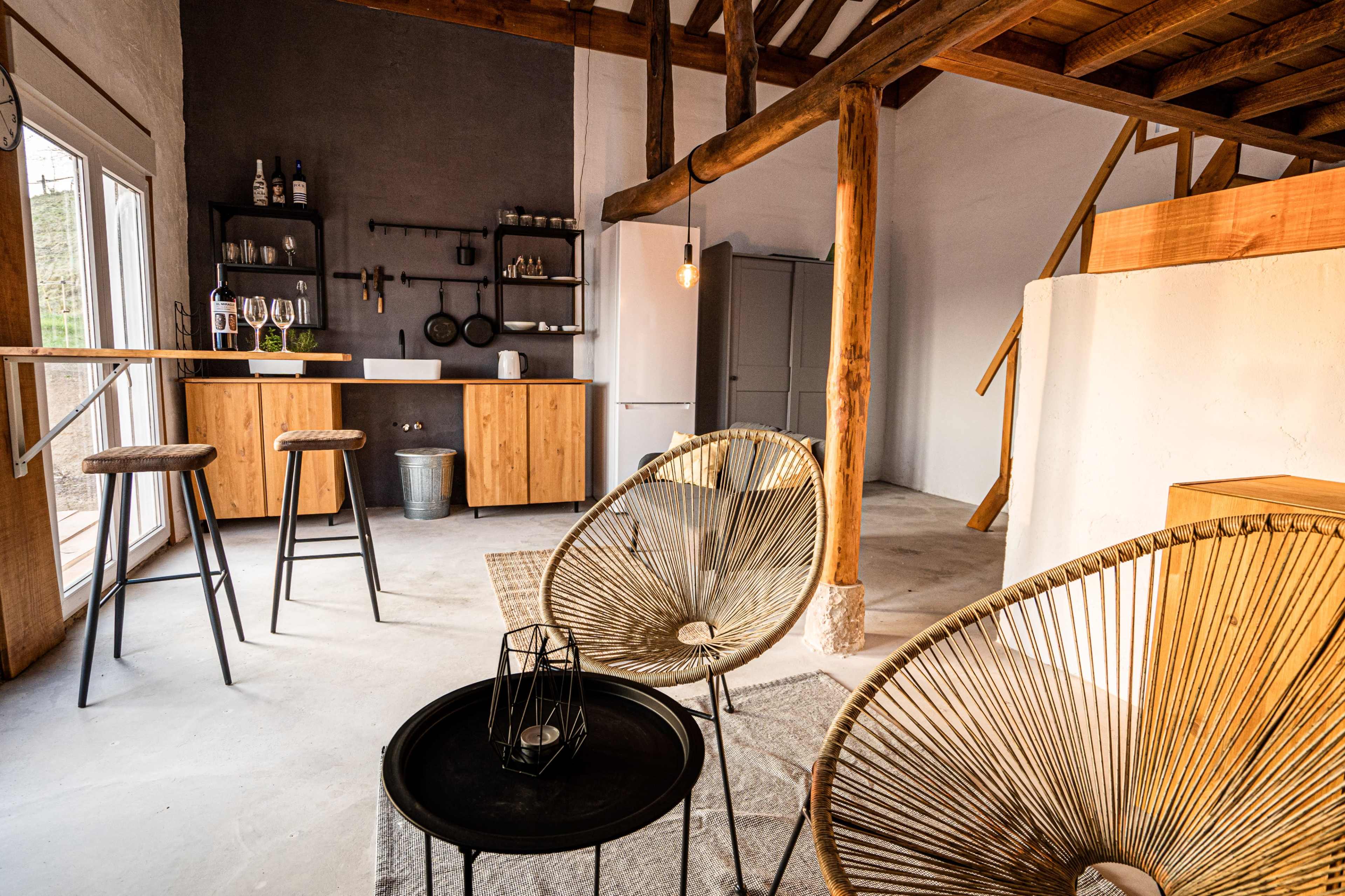 The image shows a modern, minimalist interior with wooden beams, a kitchen area, two chair designs, and a small table.