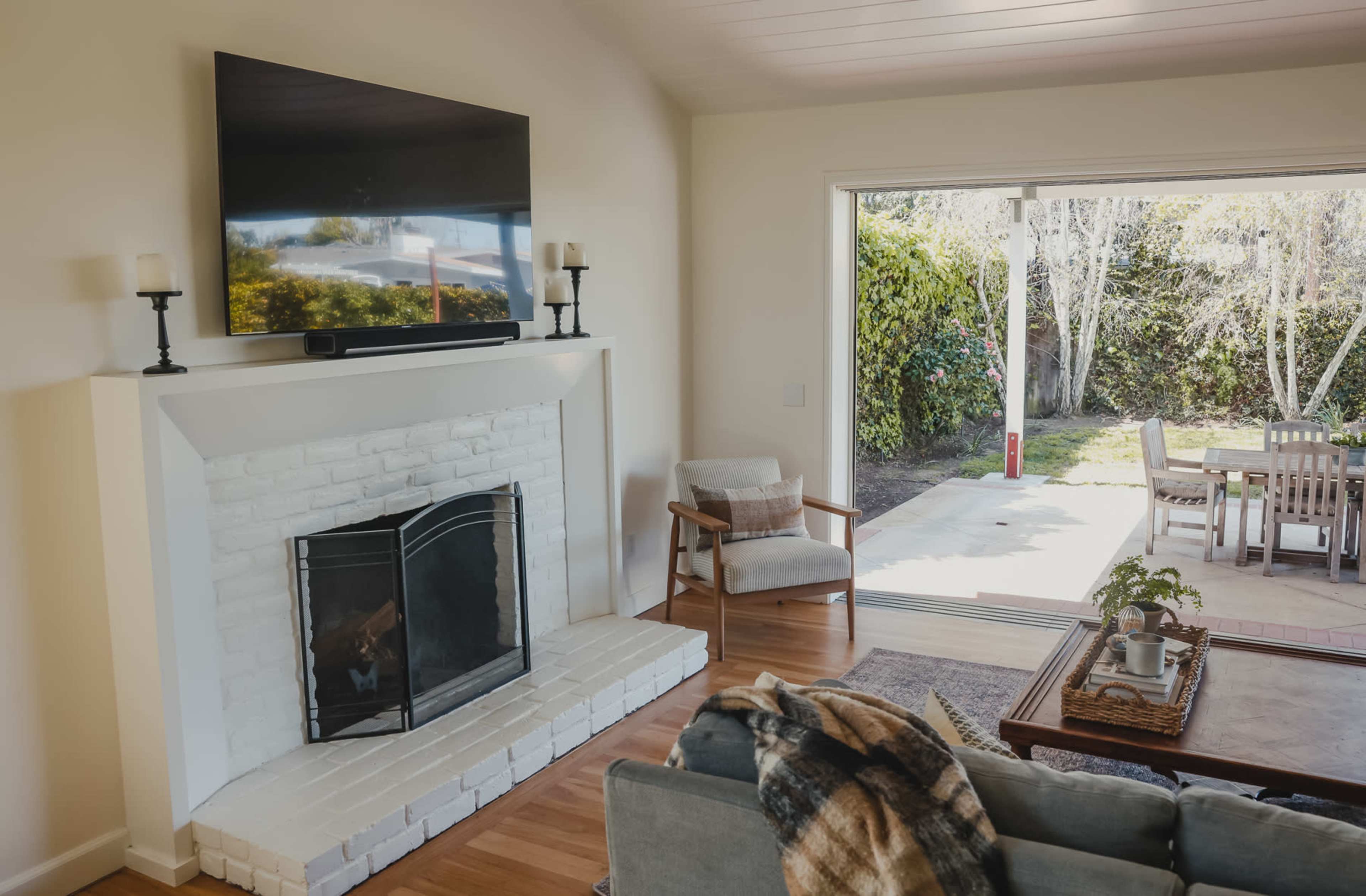 A cozy living room with a white brick fireplace and a large TV mounted above it, overlooking a patio with outdoor dining furniture.
