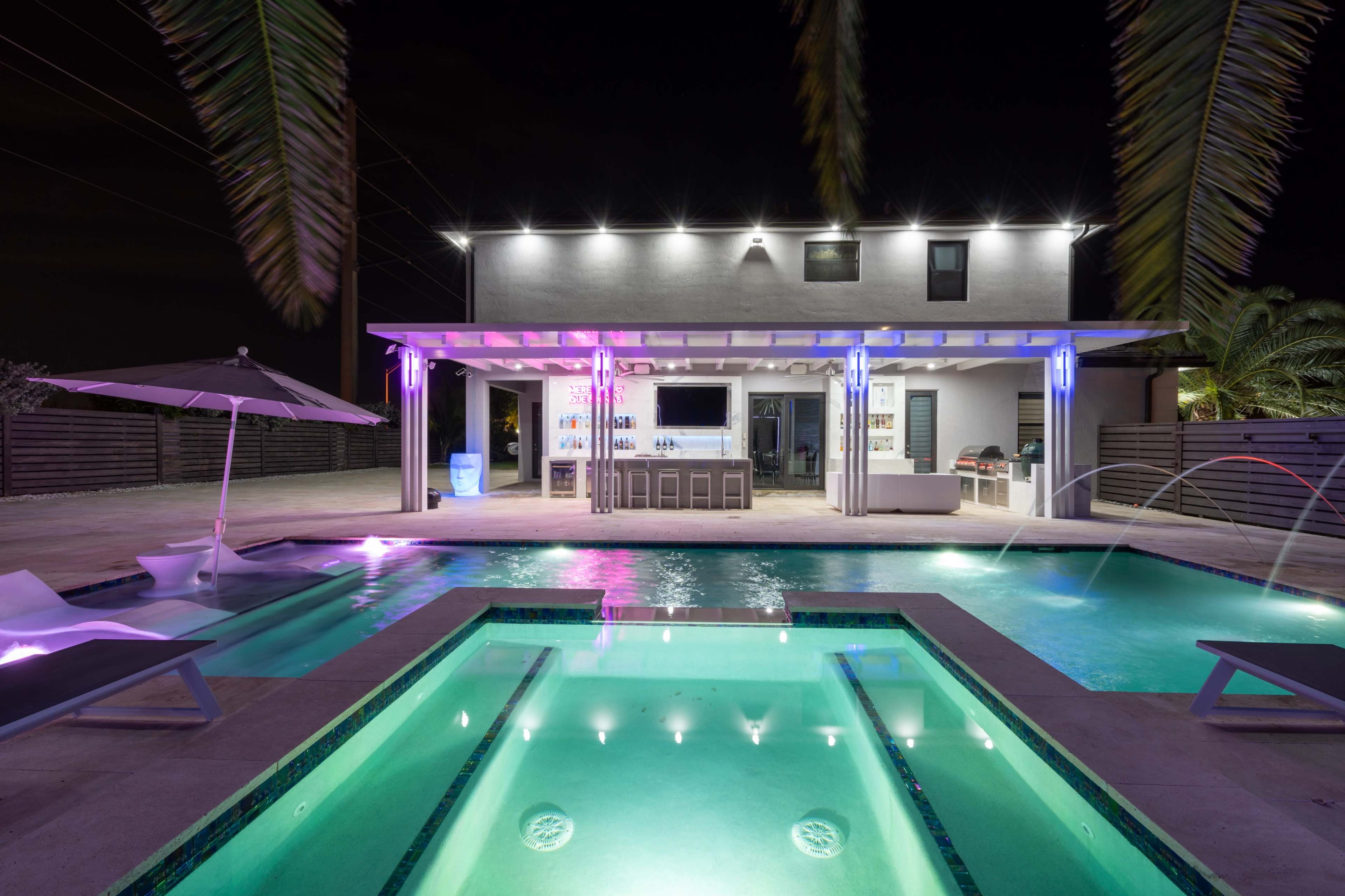 A modern outdoor pool area at night, featuring a rectangular pool with built-in lights, a bar under a pergola, and lounge chairs beside the water.