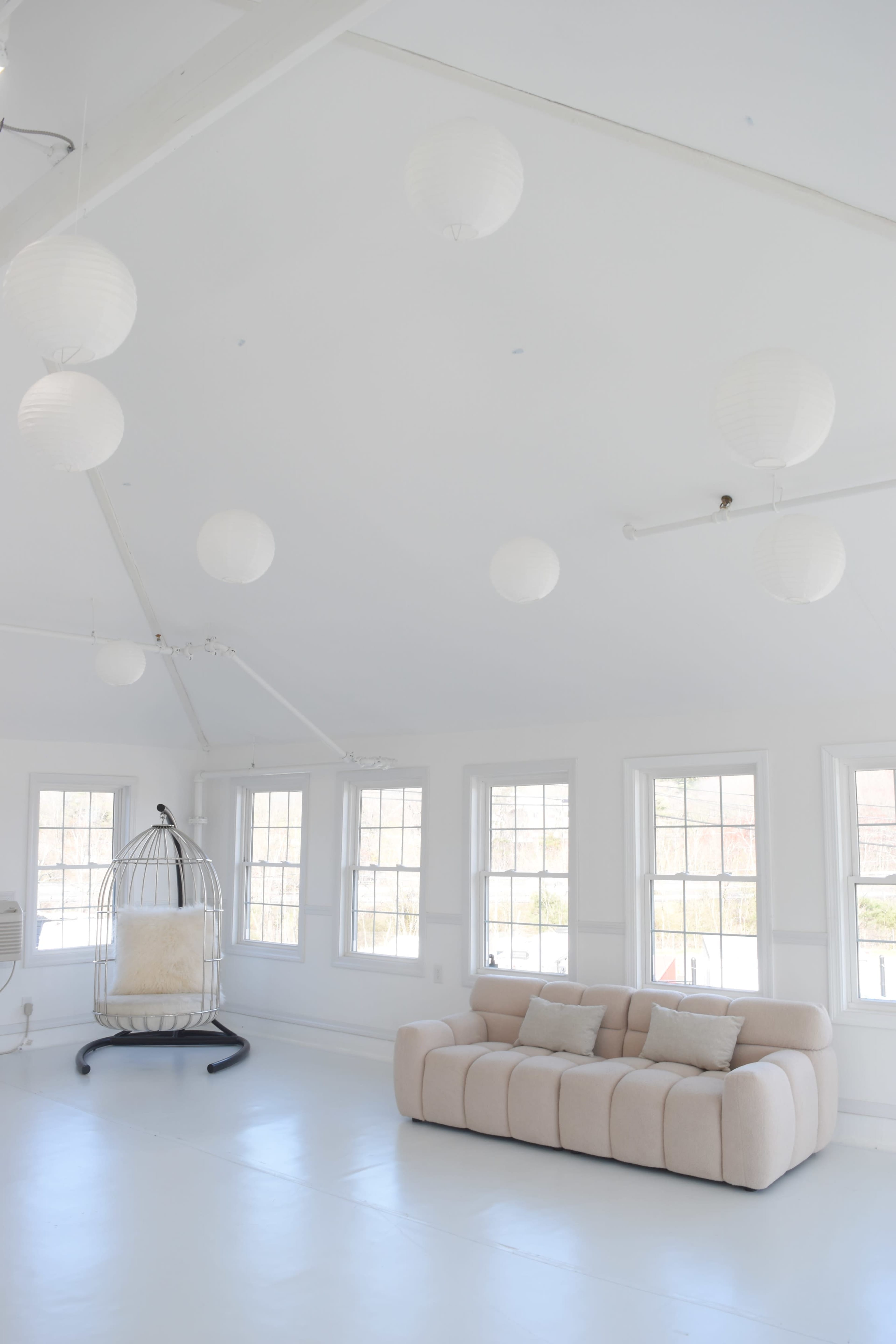 The image shows a light-filled room with white walls and ceiling, featuring a beige sofa and a hanging chair, illuminated by round paper lanterns.