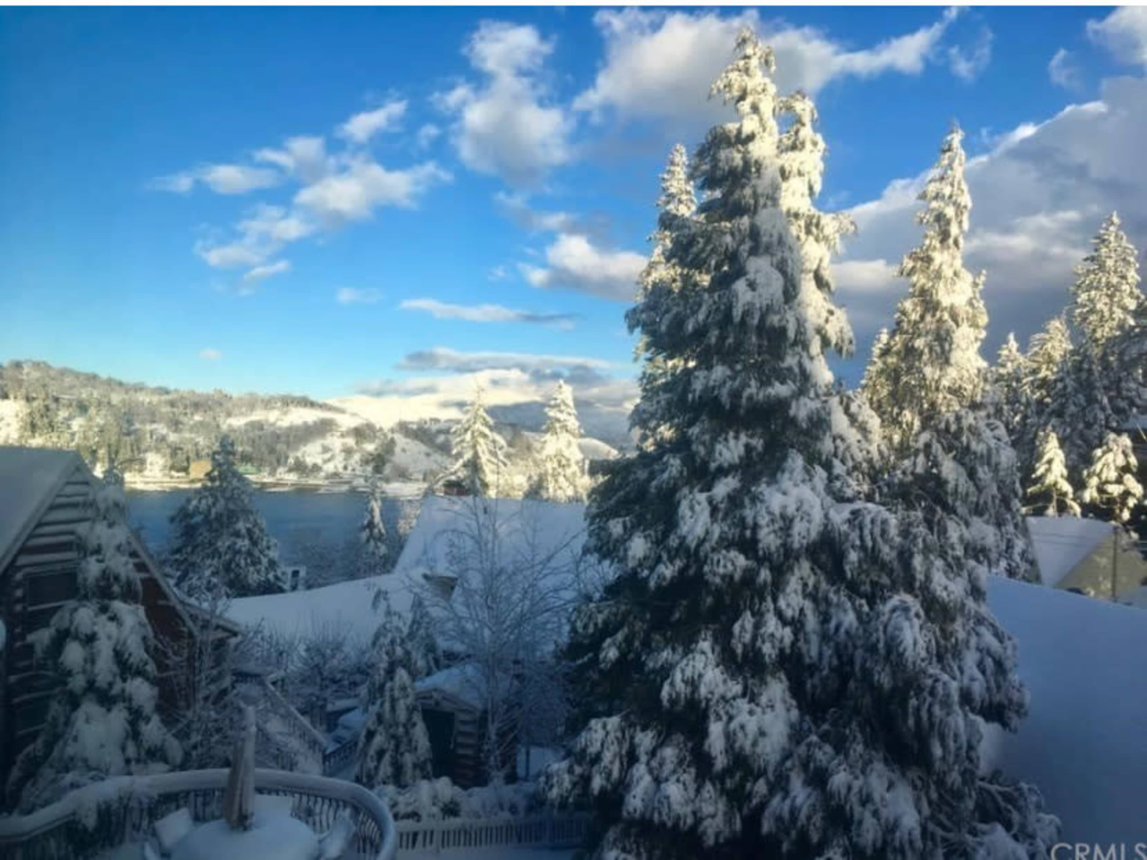 A snowy landscape features tall evergreen trees and a lake surrounded by hills under a partly cloudy sky.