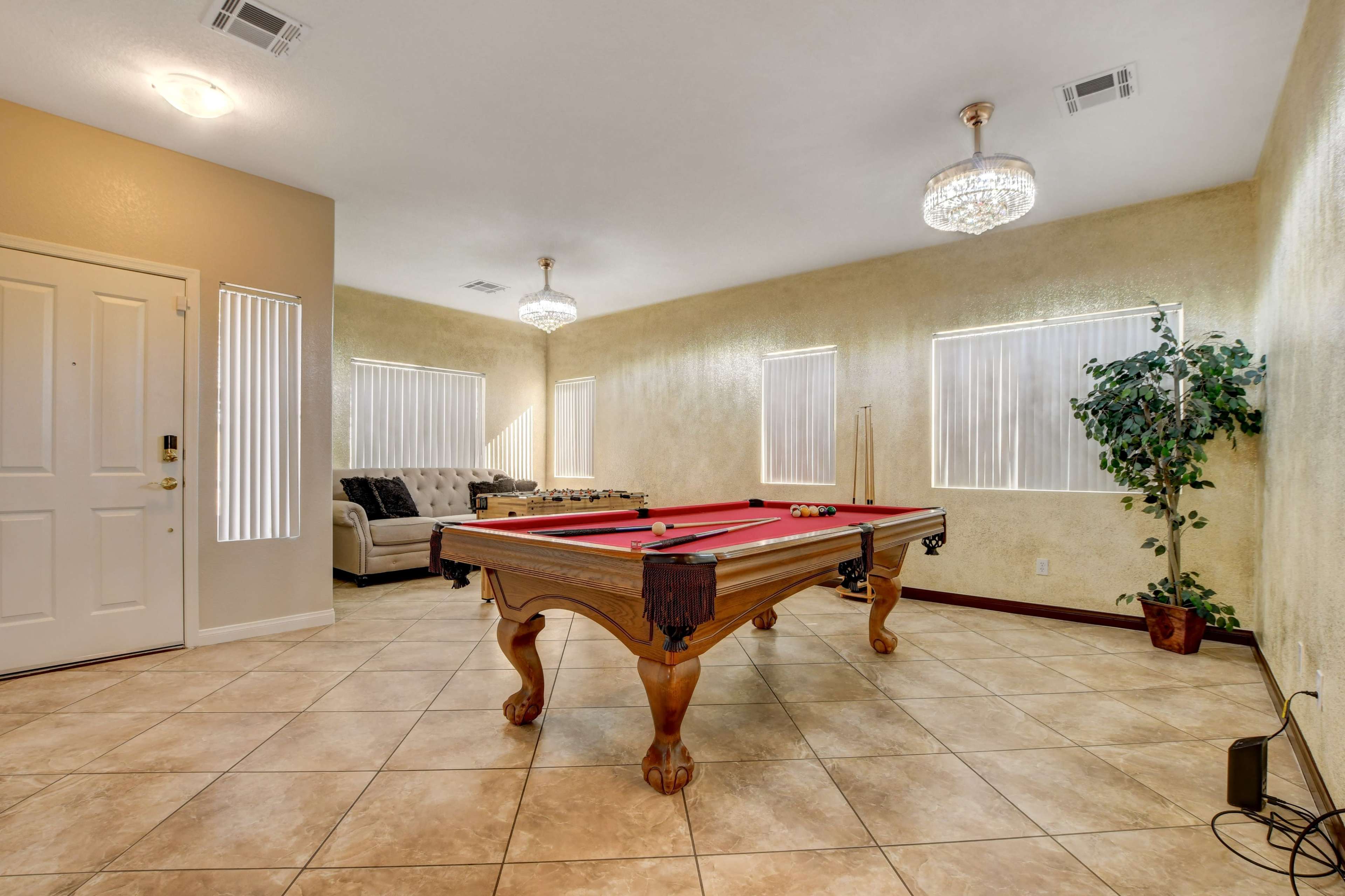 The room features a billiards table at the center, surrounded by a sofa, windows with blinds, and a decorative plant.