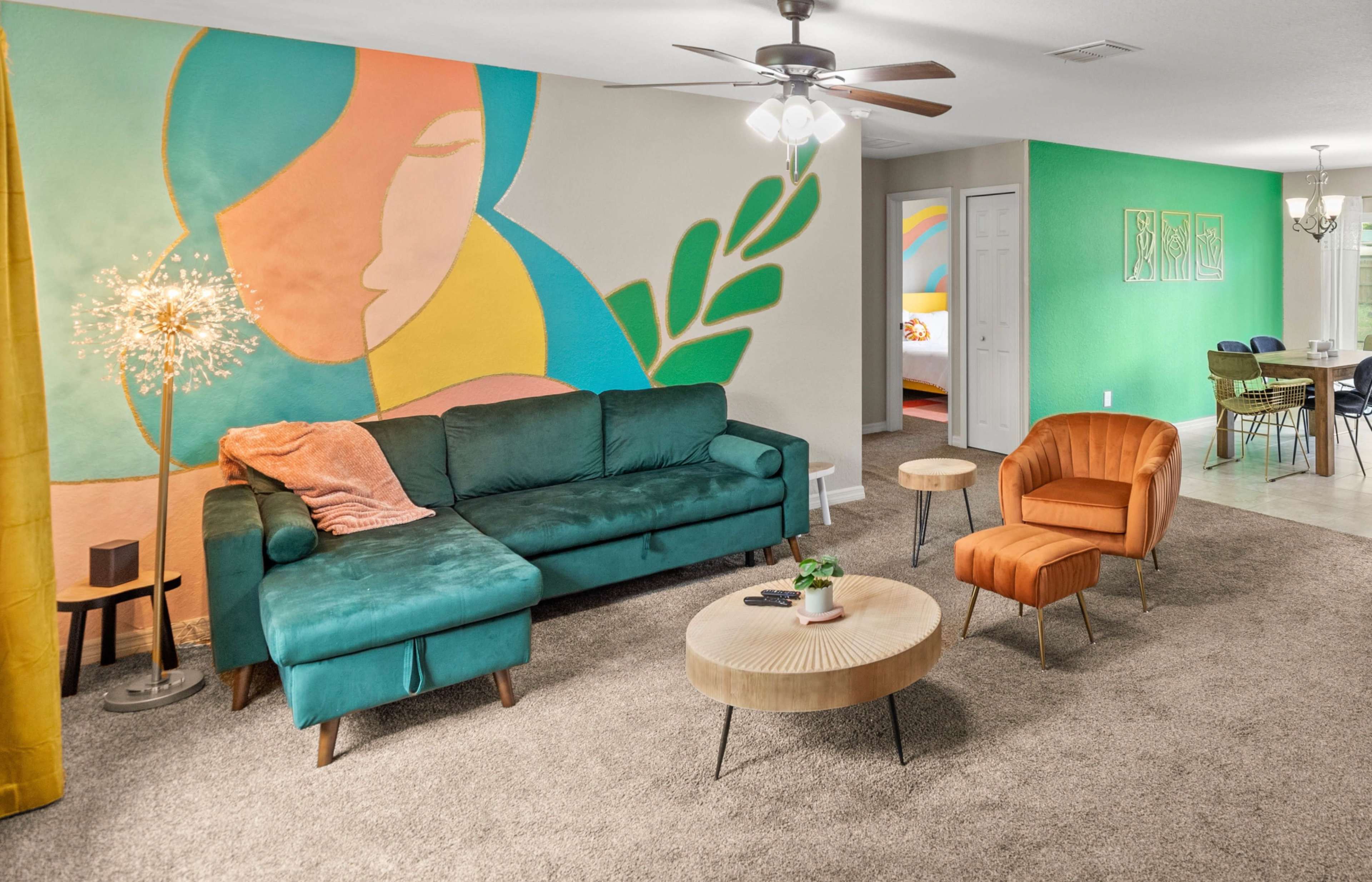 The living room features a teal sectional sofa, a round wooden coffee table, and two armchairs, alongside a colorful wall mural and a bright green accent wall.