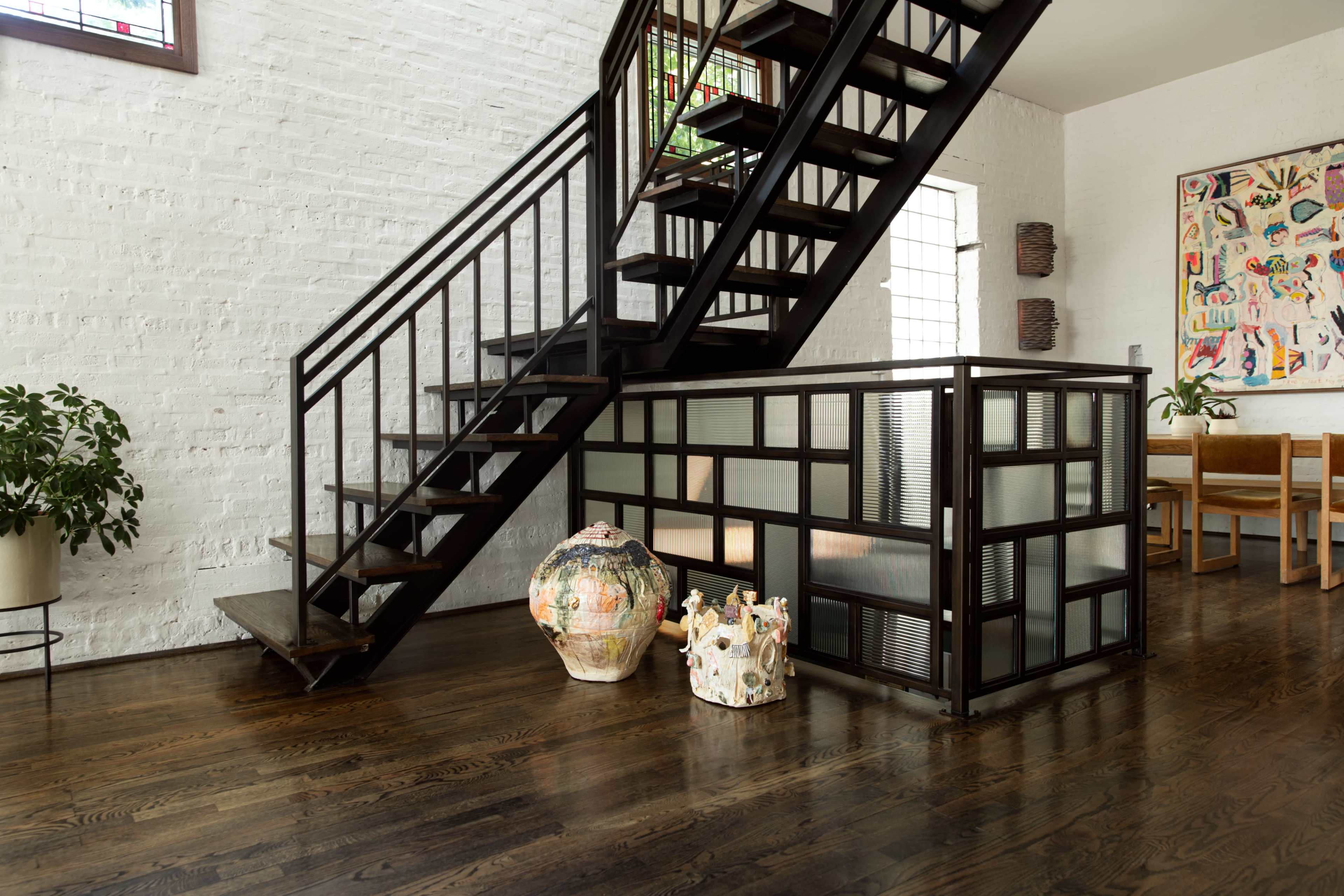 A modern interior features a metal staircase beside a glass-paneled partition, with decorative pottery placed at its base and a dining area visible in the background.
