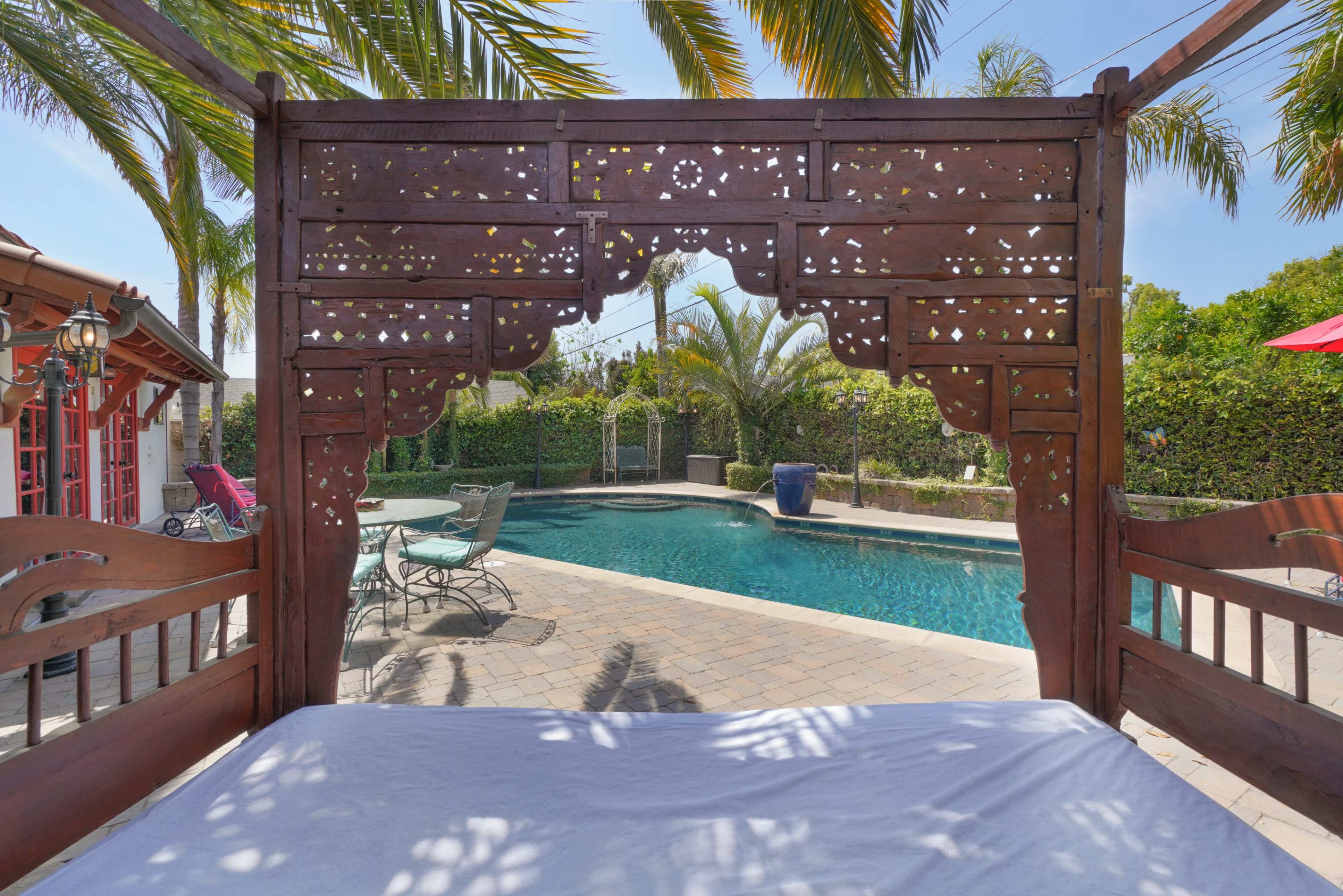 A wooden gazebo with intricate cutouts frames a view of a swimming pool surrounded by palm trees and patio furniture.